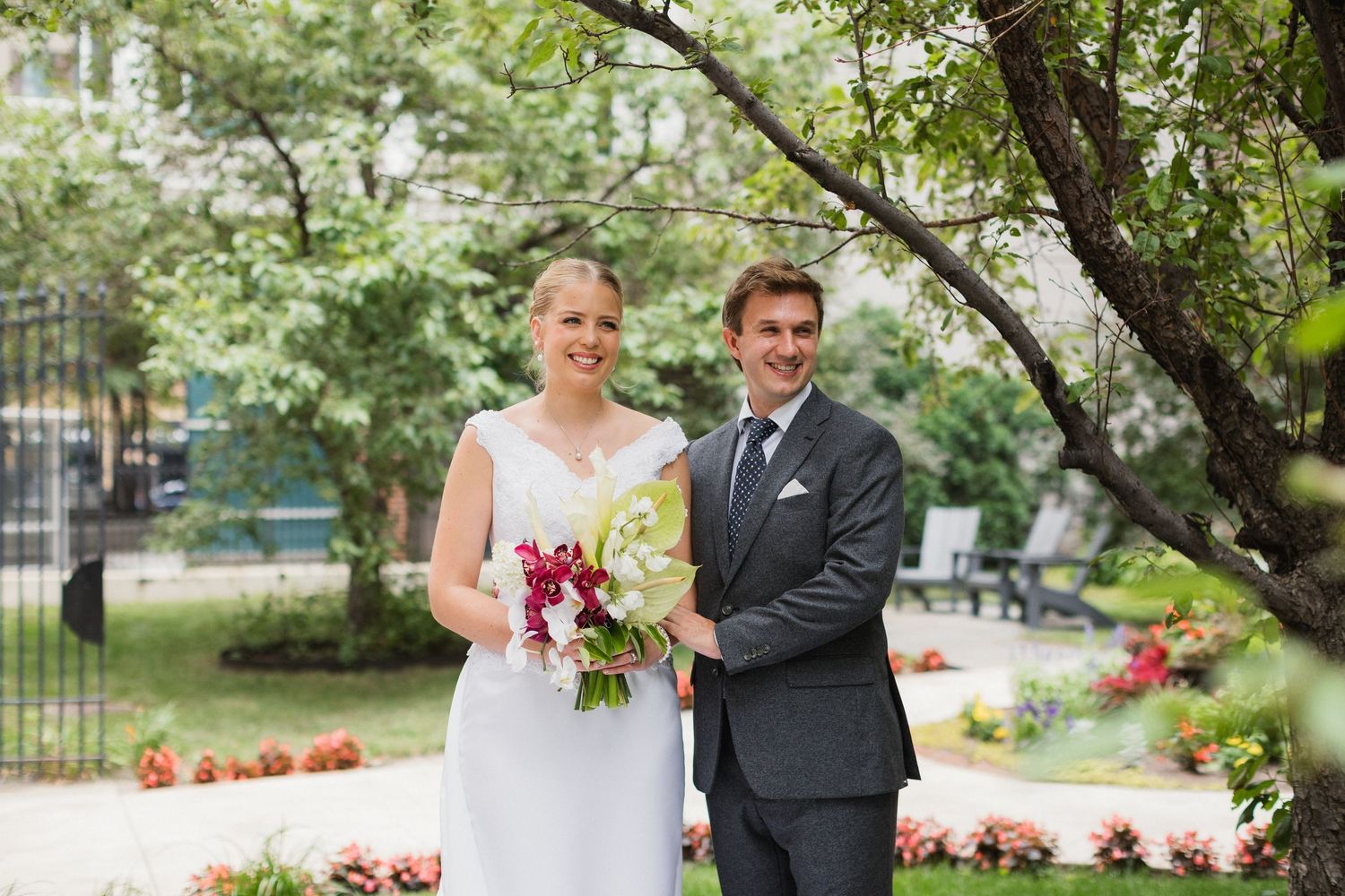 Smiling bride and groom pose together outdoors in a garden, bride holding a vibrant red and green bouquet.