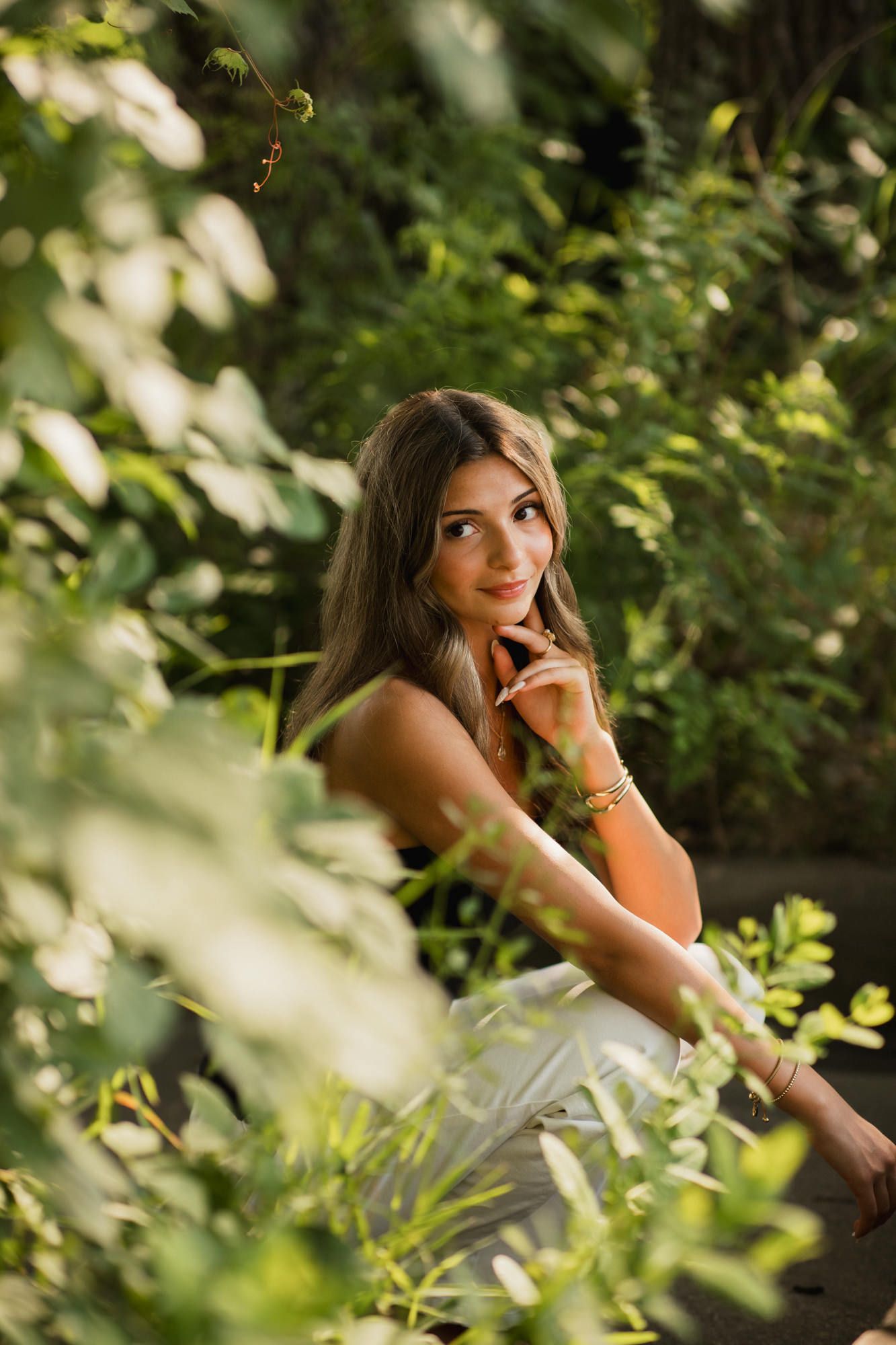 Young woman with long brown hair sits gracefully among lush green foliage, smiling softly in natural sunlight.