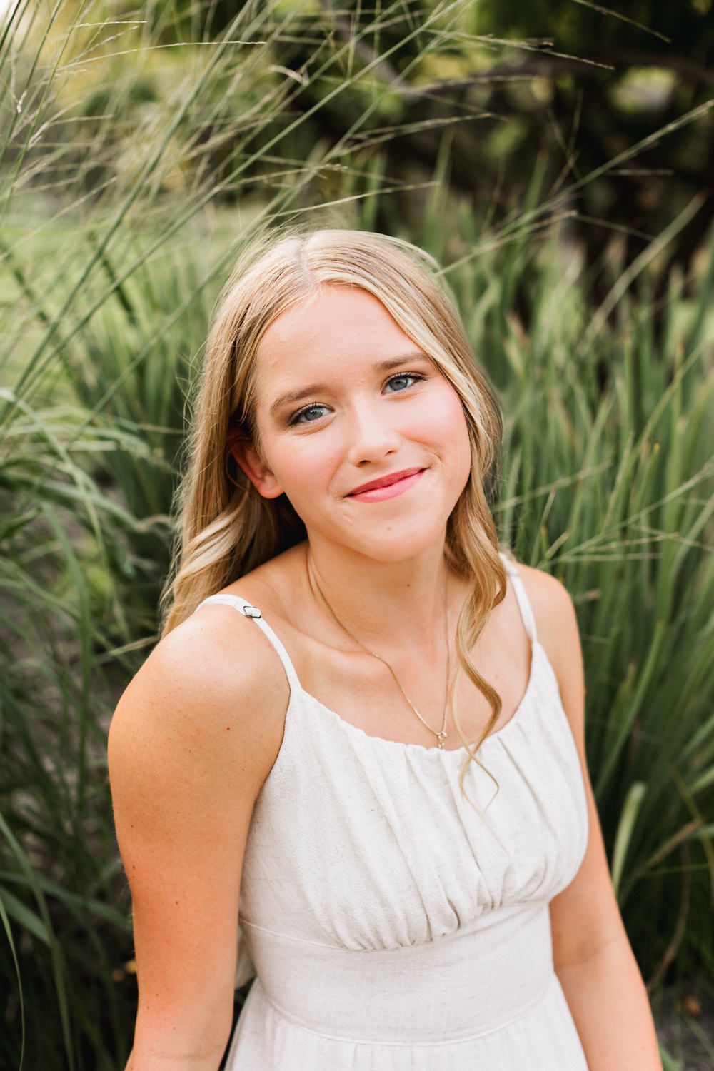 Young person in white summer dress poses against natural outdoor greenery backdrop.