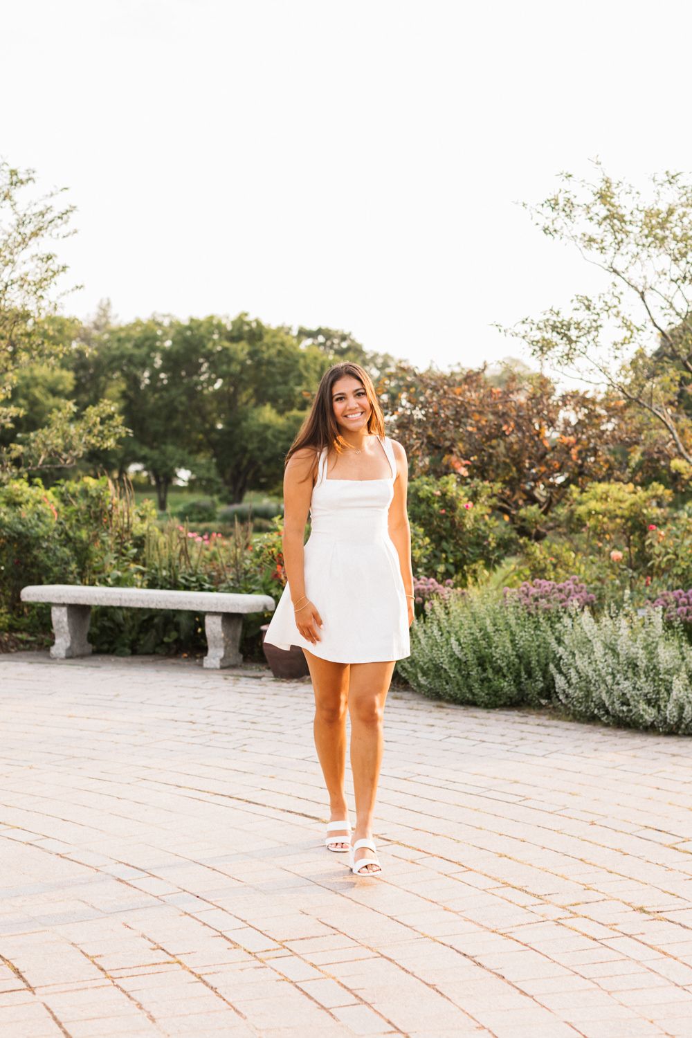 Someone in a white summer dress stands on a brick path in a manicured garden setting.