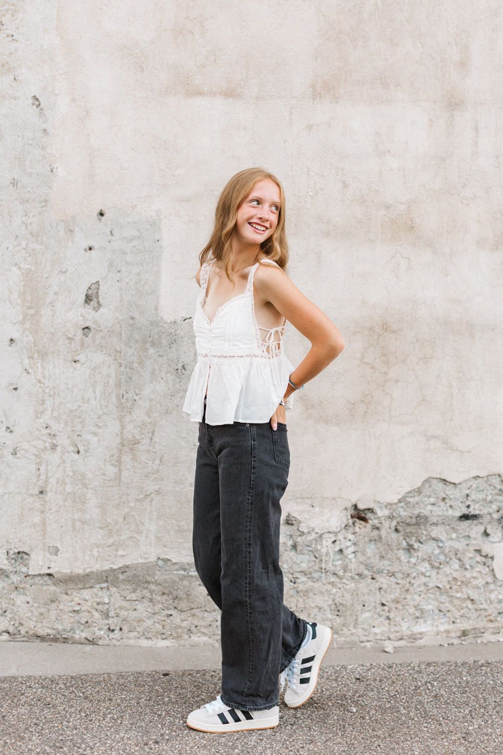 A person in a white sleeveless top and wide-leg jeans poses against a concrete wall while wearing Adidas sneakers.
