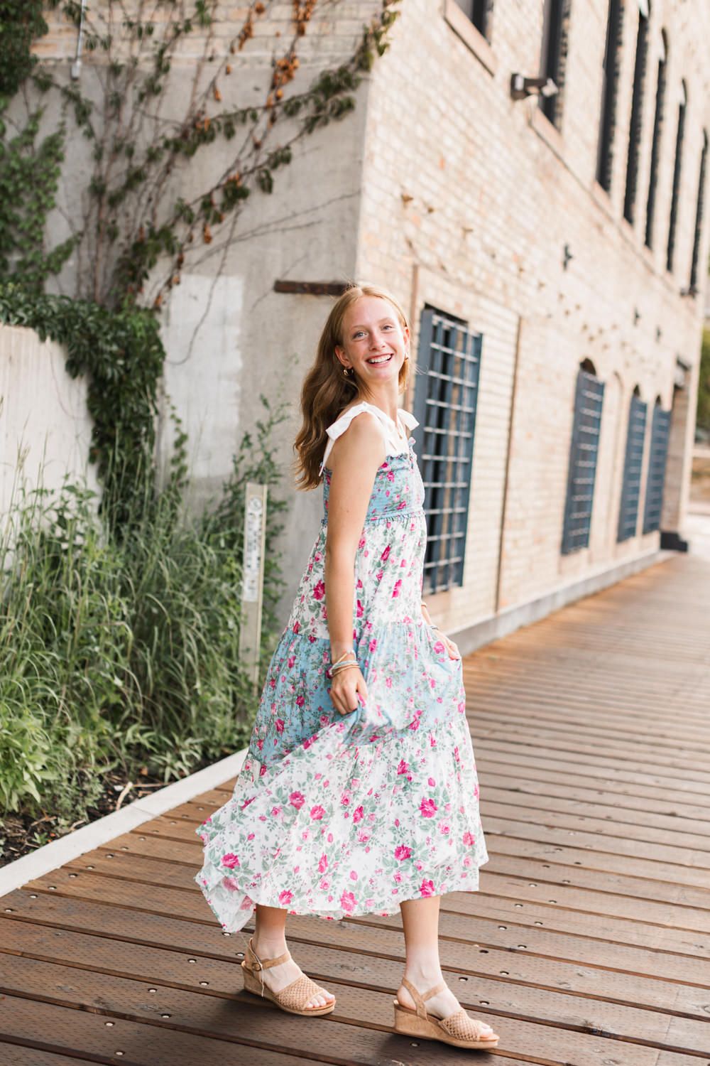 A floral summer dress twirls on a wooden deck with brick building and greenery in background.
