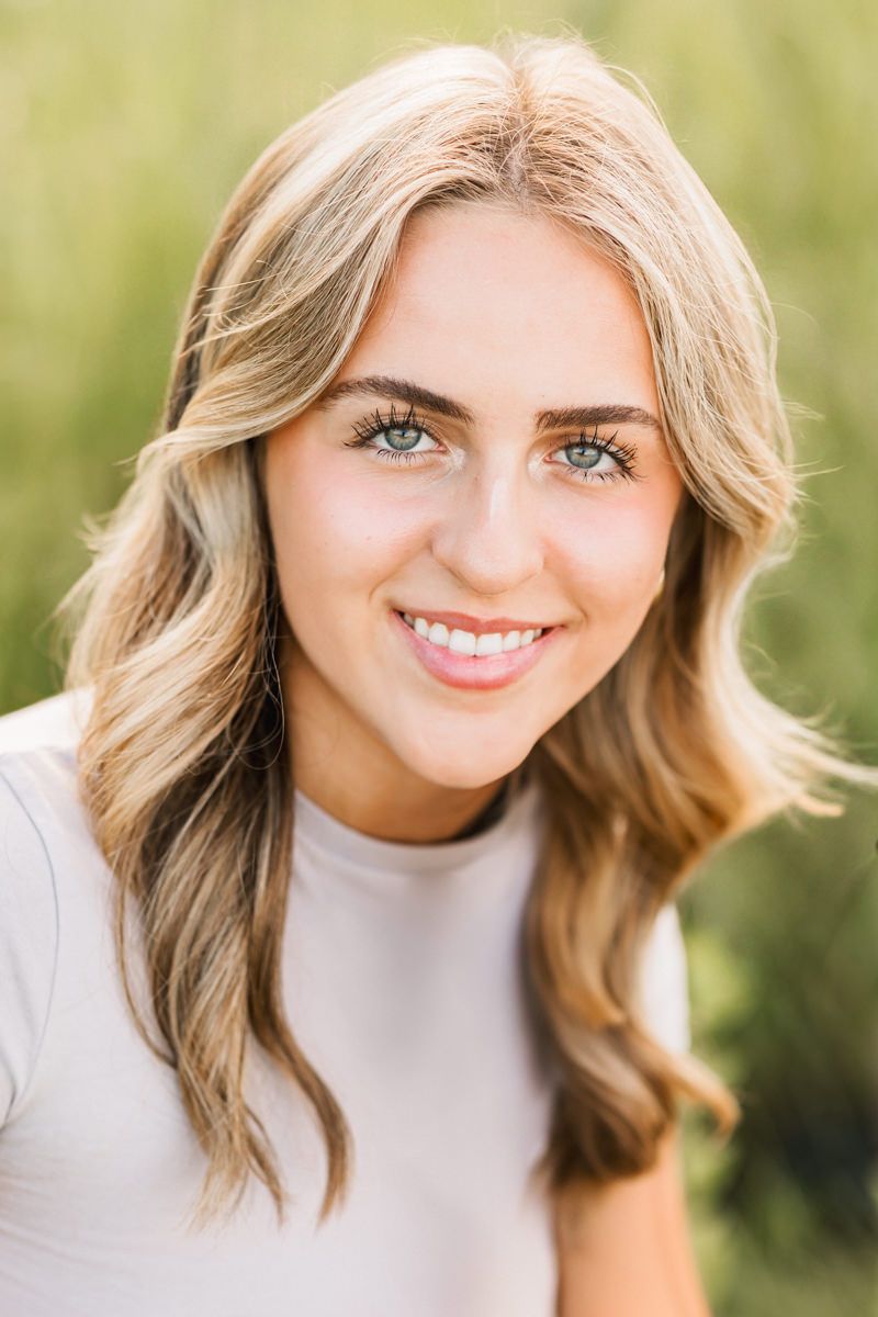 Portrait of a person with blonde wavy hair and bright blue eyes wearing a light colored top against a blurred green background.