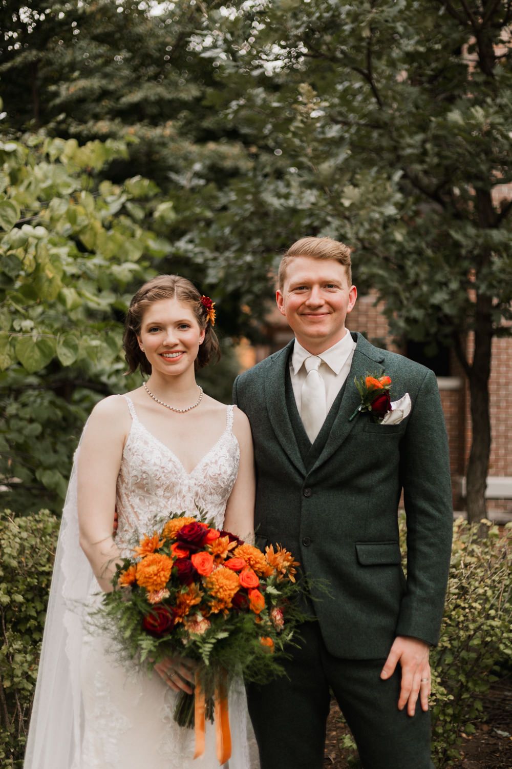 Newlywed couple poses outdoors with vibrant orange and red autumn bouquet against green foliage background.
