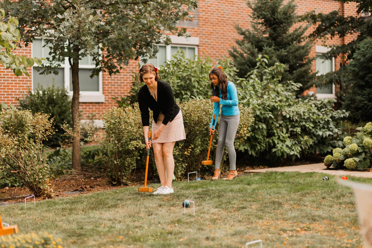 A panoramic view of people playing fetch with a dog on a sunny day in a residential backyard with brick buildings.