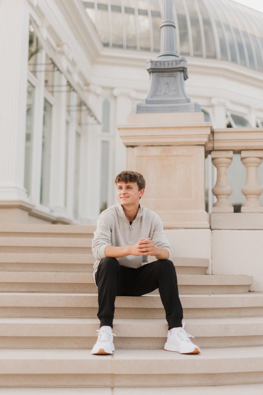 Someone sits contemplatively on light stone steps wearing casual attire and white sneakers.