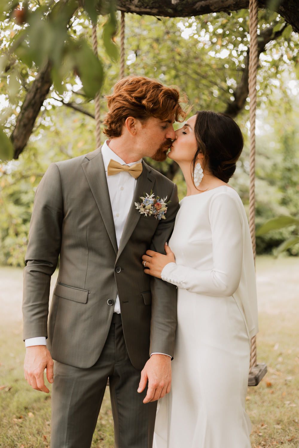 A romantic wedding couple share a tender moment on a rustic rope swing in a sunlit garden setting.