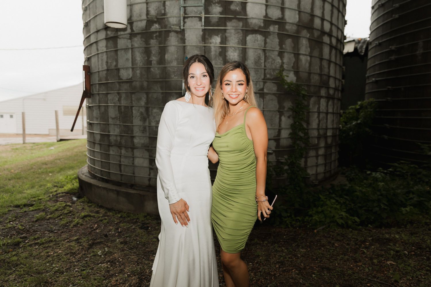 Two people pose together against large dark silos in an outdoor setting.