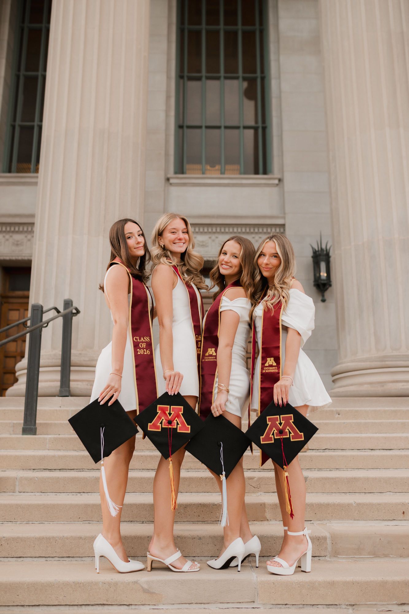 Four female University of Minnesota graduates in white dresses hold maroon M graduation caps on steps.