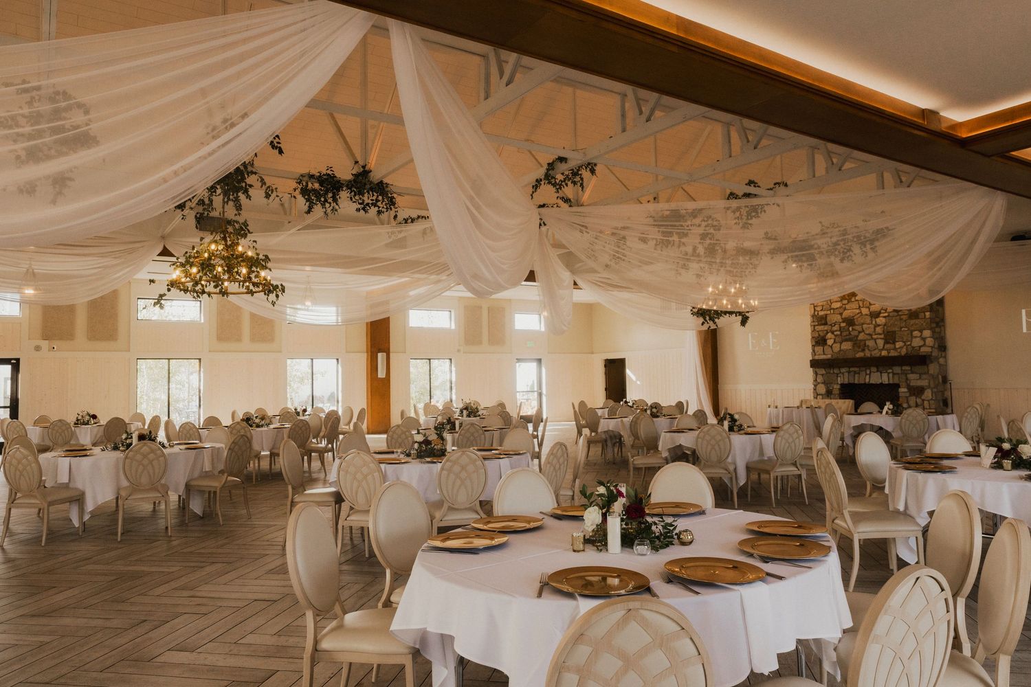 Elegant wedding reception room with round tables, draped white fabric, chandeliers and a stone fireplace ready for guests.