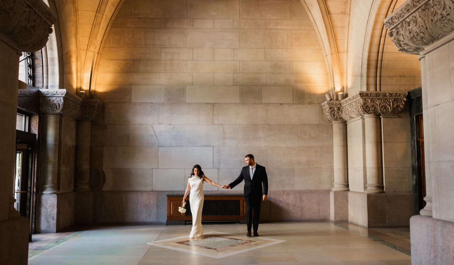 Elegant couple holding hands in a grand stone archway hall, bride in white pantsuit, groom in dark suit.
