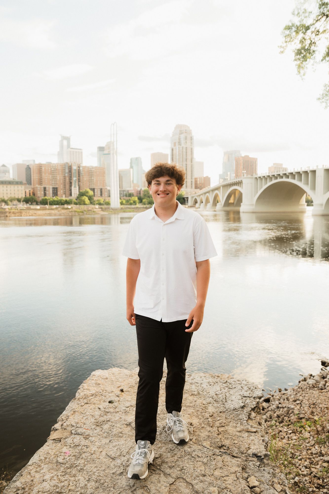 Young man in white shirt smiling by Mississippi River with Minneapolis skyline and bridge in background.