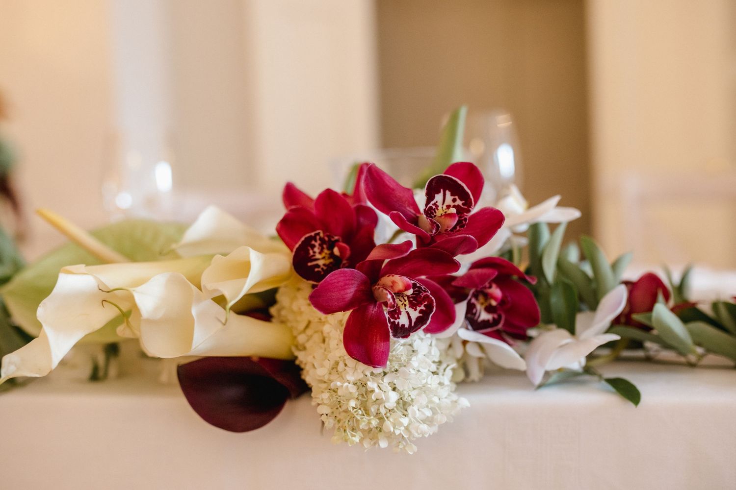 Elegant wedding centerpiece with deep red cymbidium orchids, white calla lilies, and baby's breath on a white tablecloth.