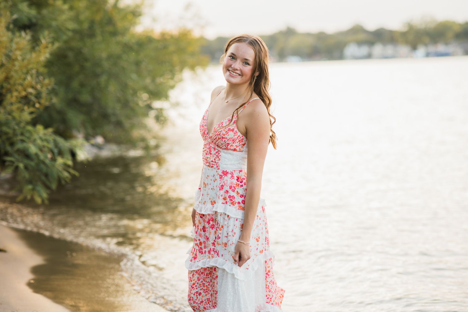Young woman in floral dress smiling on lakeside beach surrounded by lush green trees in warm golden light.