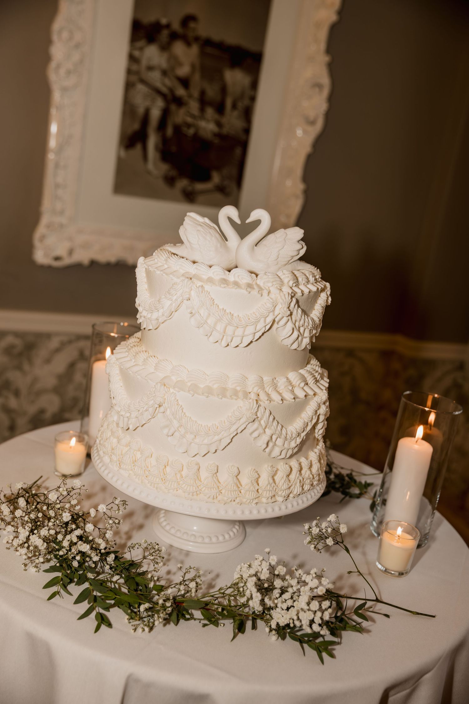 Three-tier white wedding cake with swan toppers, piped frosting details, candles, and baby's breath floral arrangement.