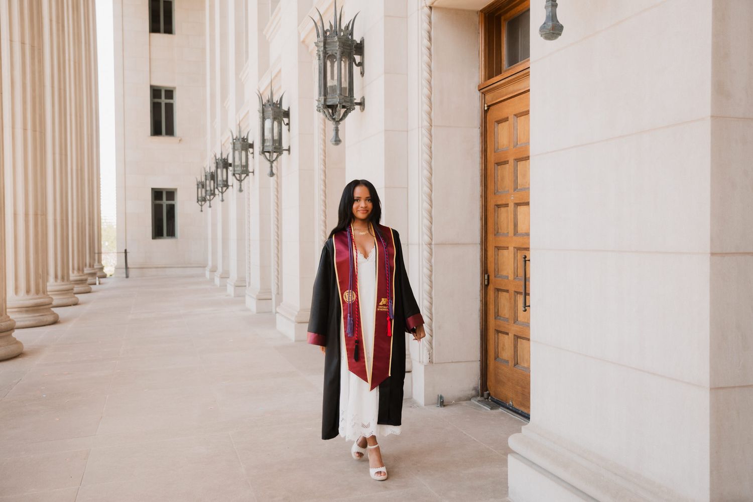 A graduate in black and maroon regalia walks confidently between grand columns at a university building.