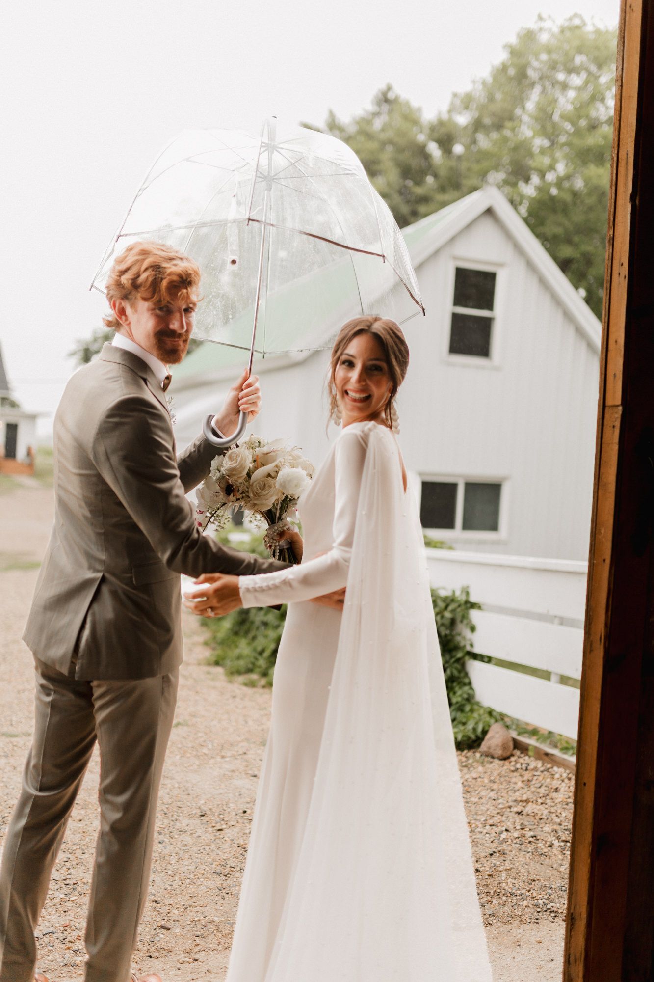 Bride in white cape gown and groom in gray suit share a clear umbrella while holding a bouquet outside a barn.