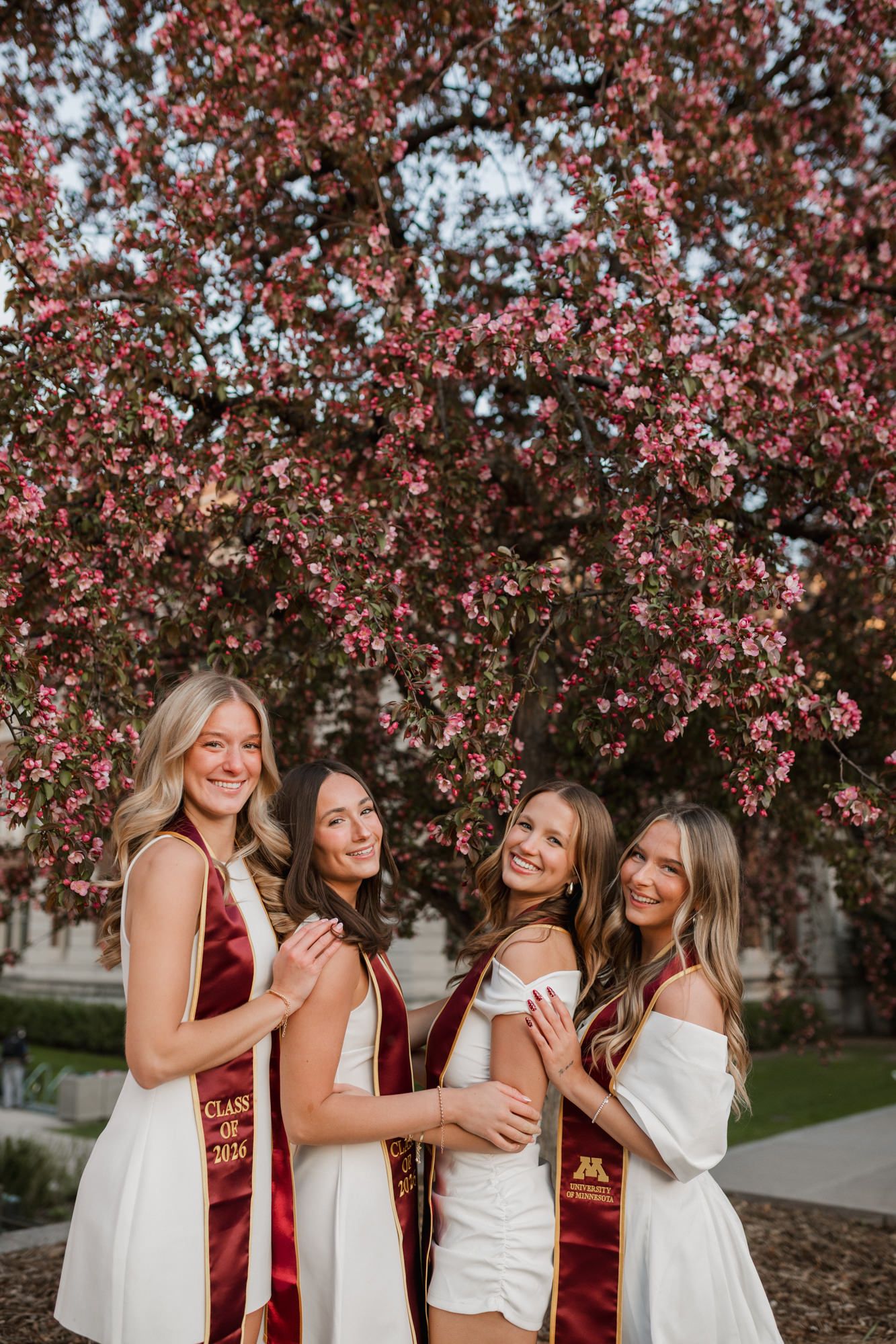 Four smiling women in white dresses and maroon graduation stoles pose under a blooming pink flowering tree.