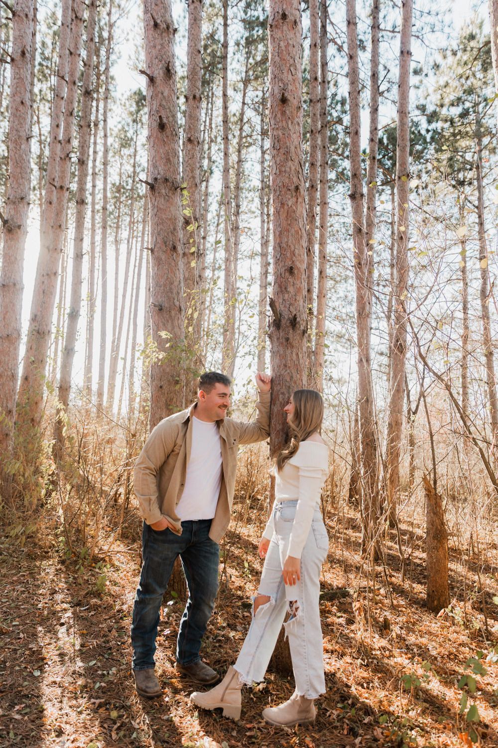 A couple in casual attire shares romantic moments among tall pine trees on a sunny autumn day in the forest.