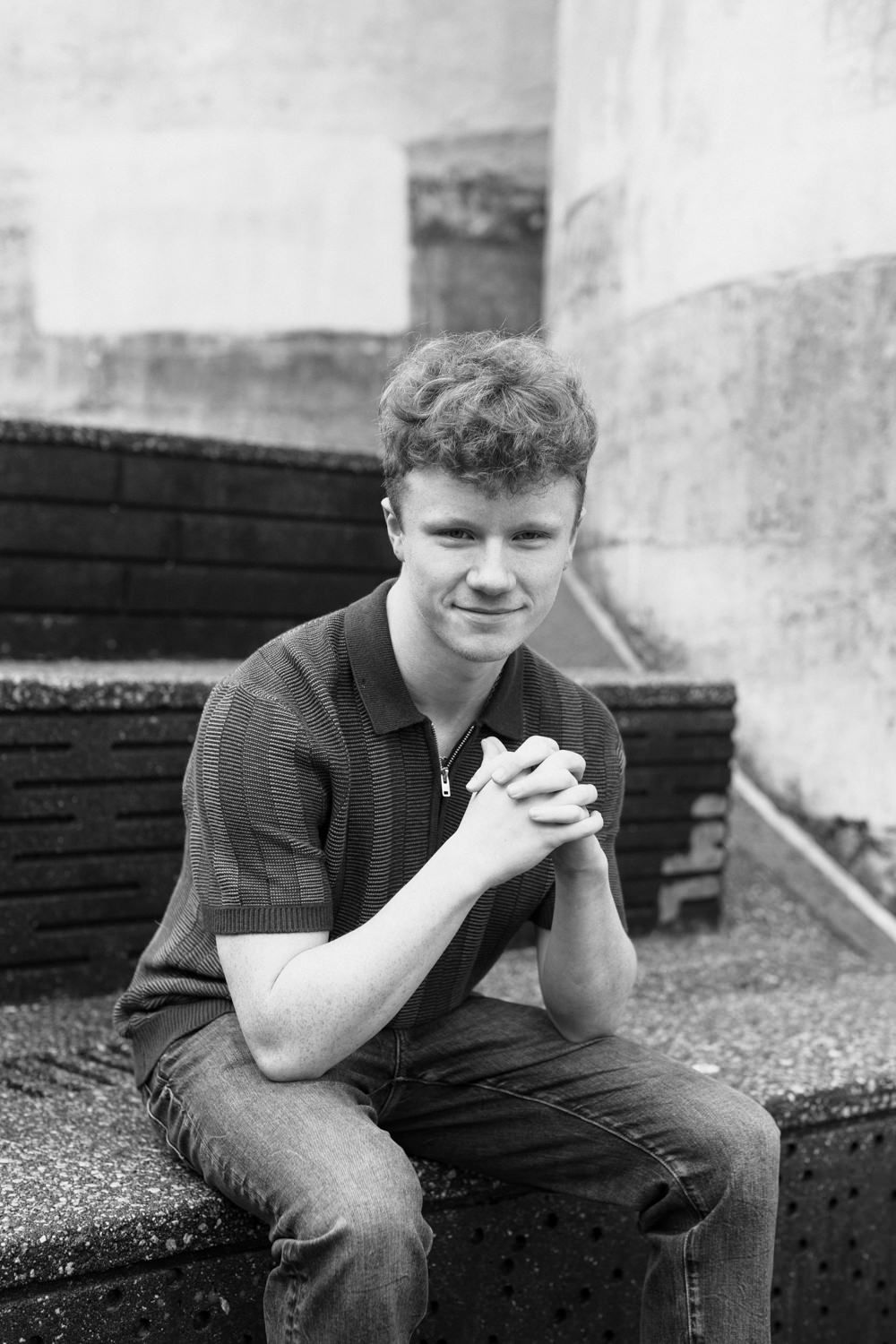 Black and white portrait photo of a teen sitting casually on a bench against a textured wall.