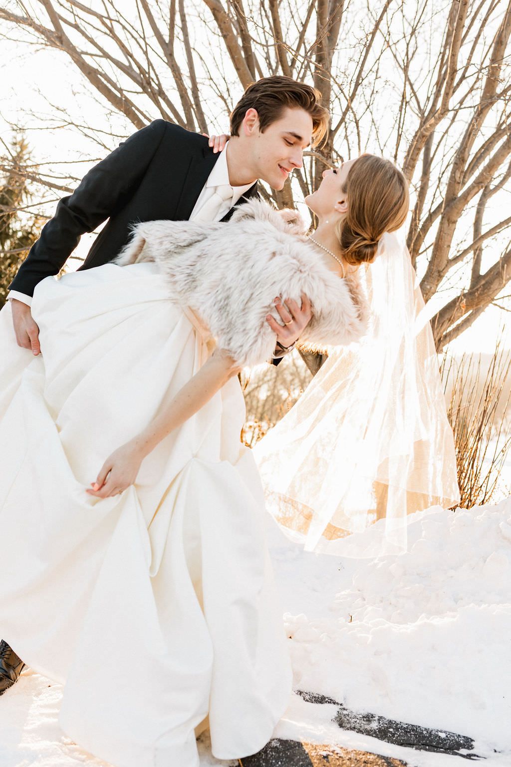 Wedding couple dance and twirl together in snowy winter scene with bare trees in background.