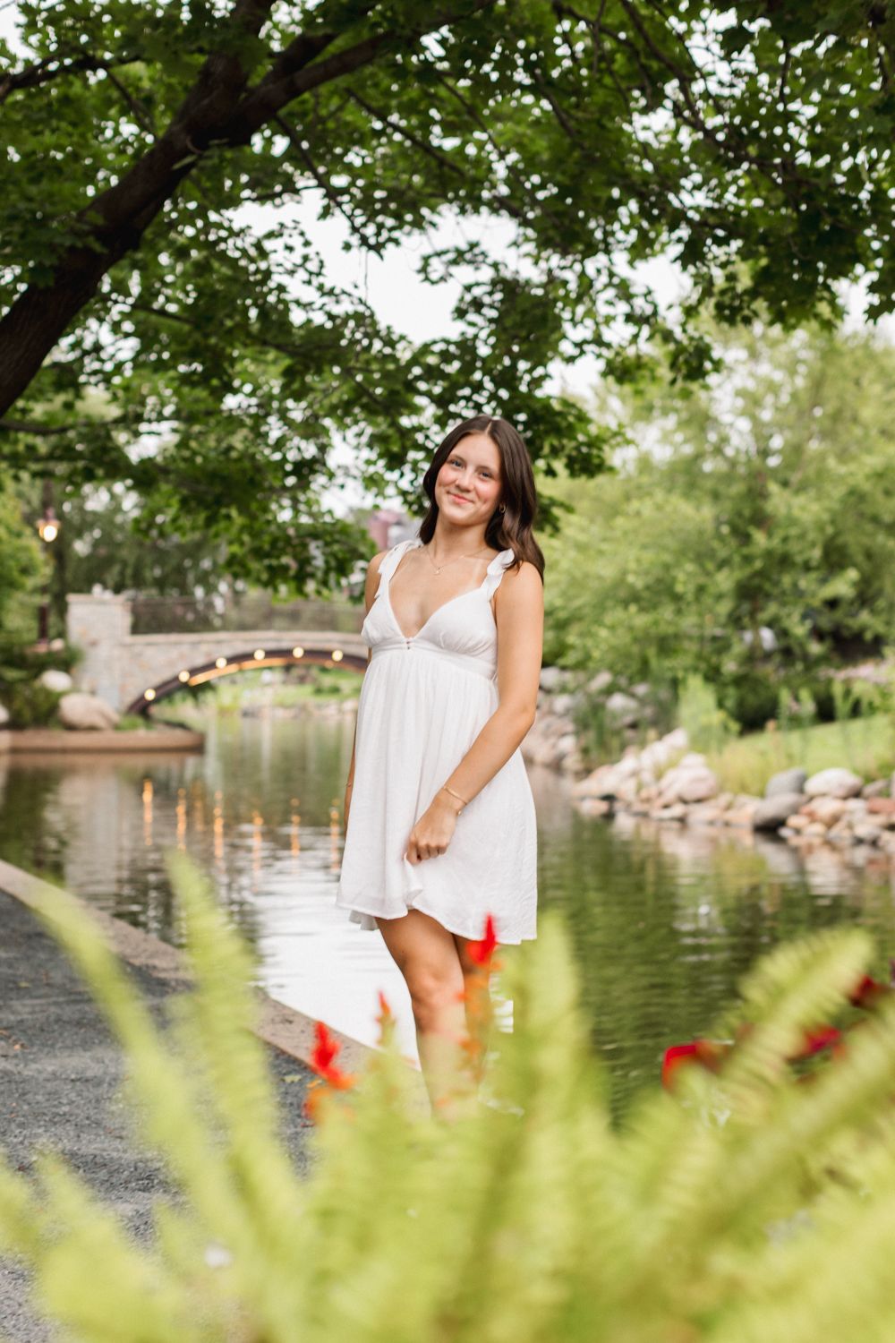 A person in a flowing white sundress stands by a tranquil pond with green trees and landscaping in the background.
