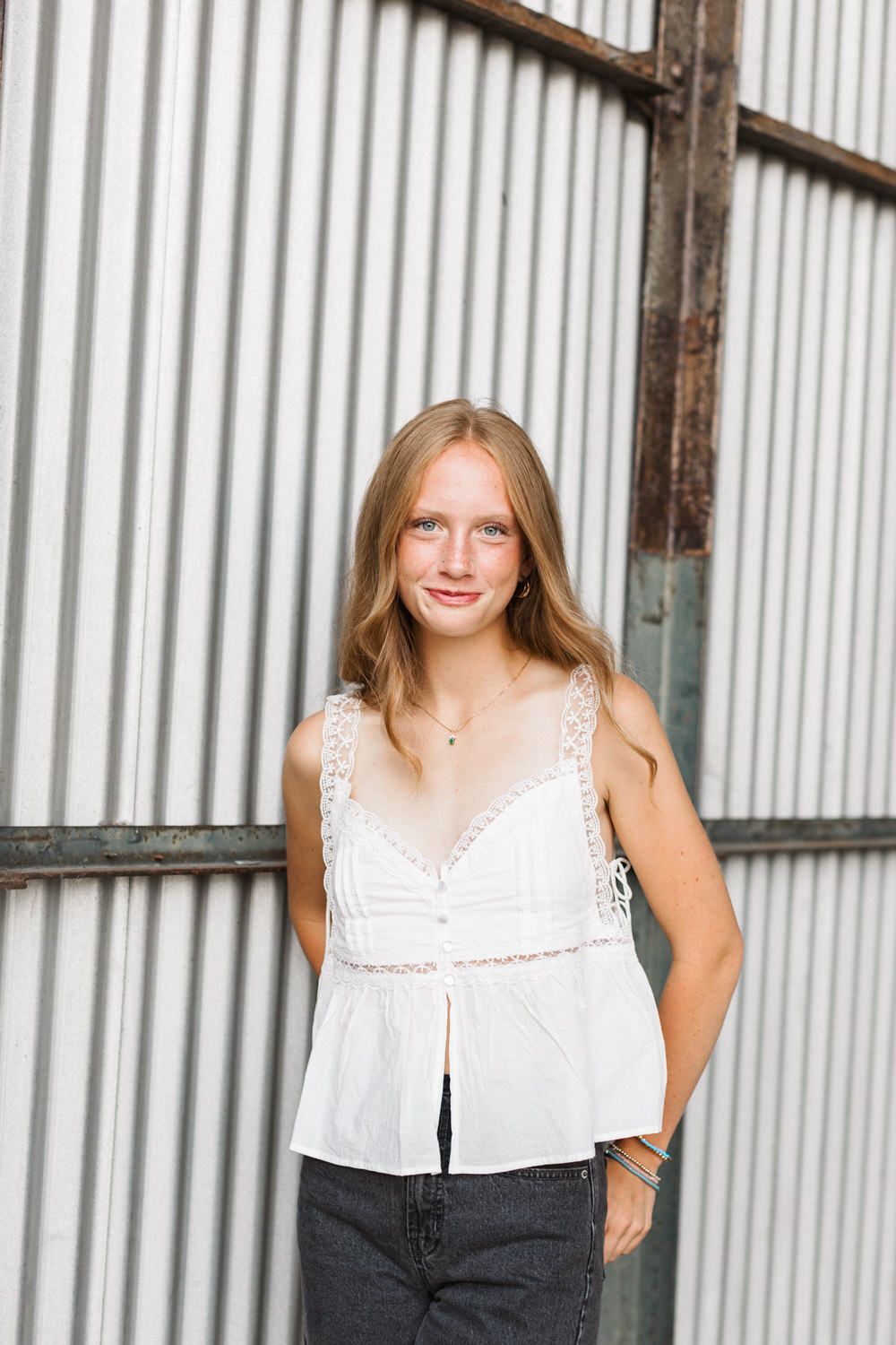A relaxed portrait by a corrugated metal wall shows someone wearing a white lace-trimmed top and dark jeans.