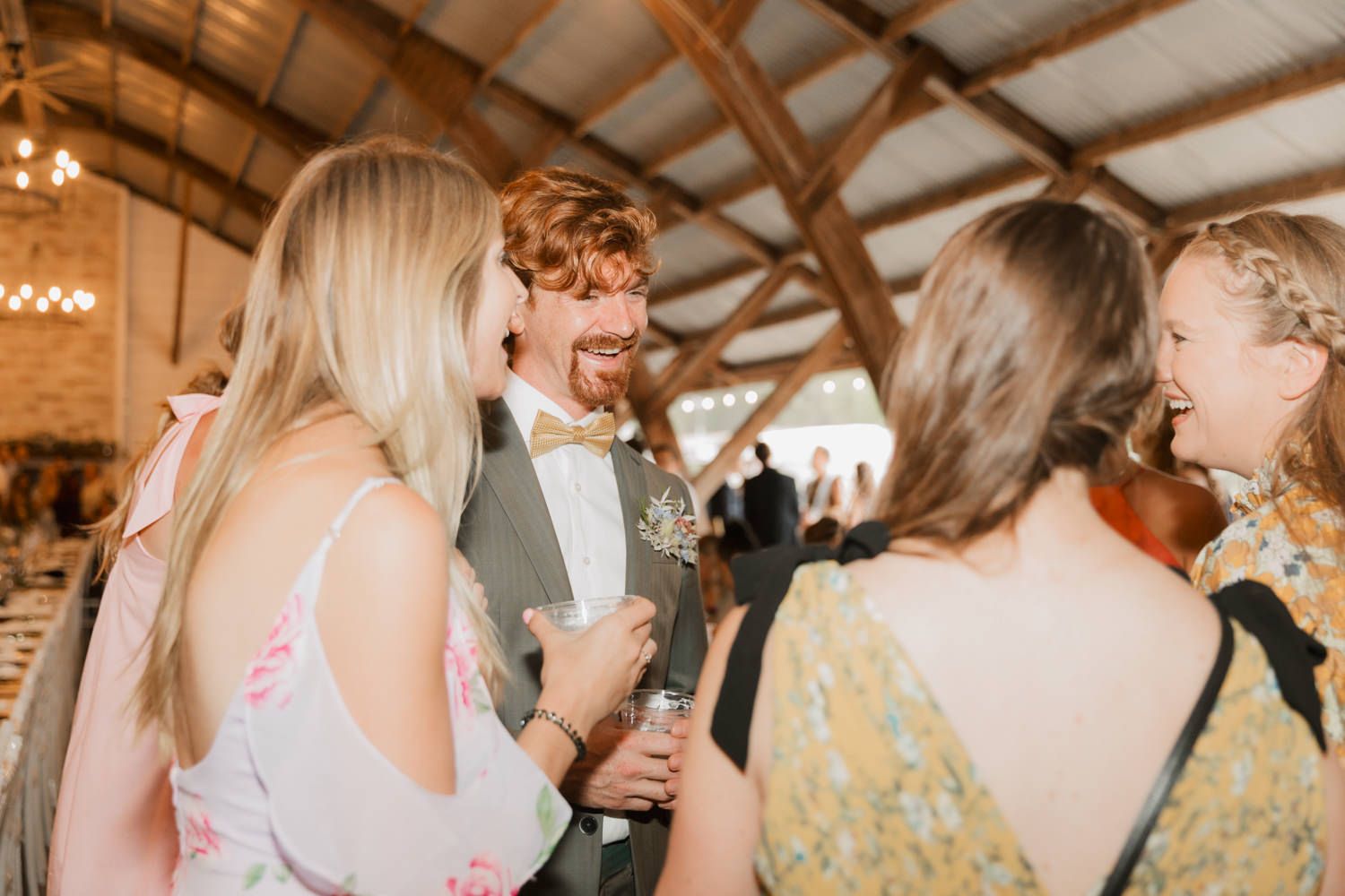 Wedding guests share laughter and conversation at a rustic barn reception venue with warm lighting and wooden beams.