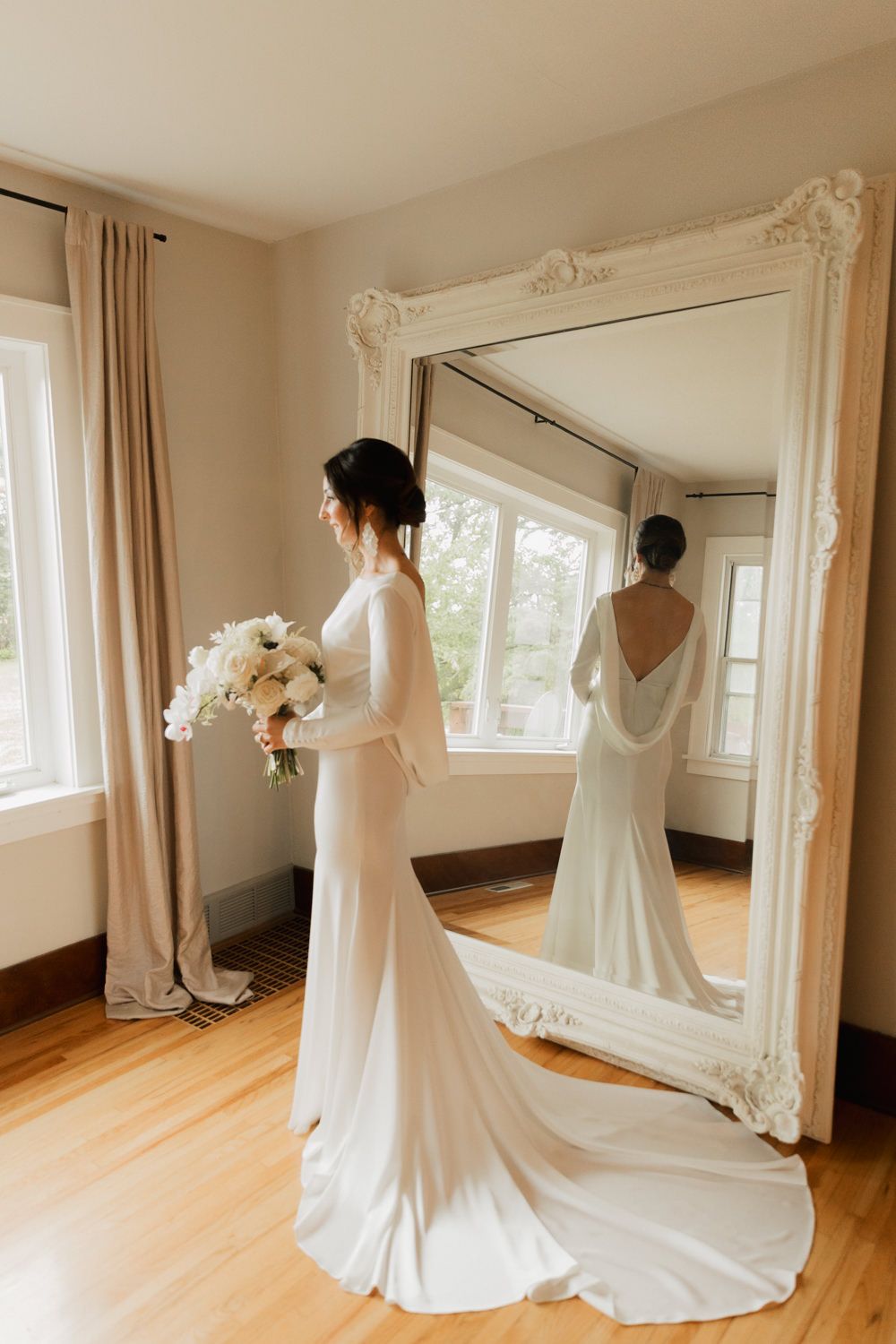 Elegant white wedding dress with flowing train displayed in a bright room with hardwood floors and natural lighting.