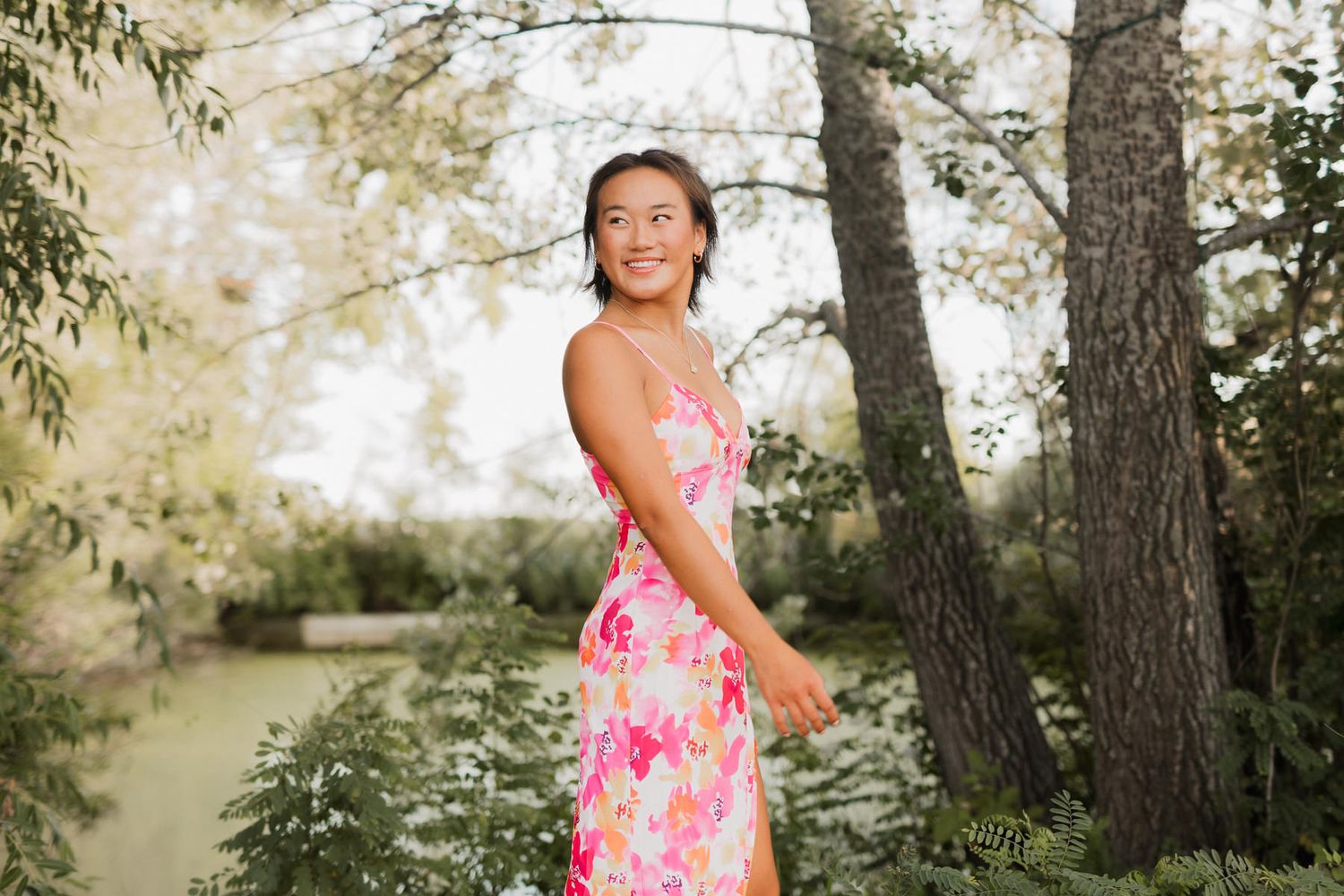Young woman in pink floral dress smiling outdoors among lush green trees.