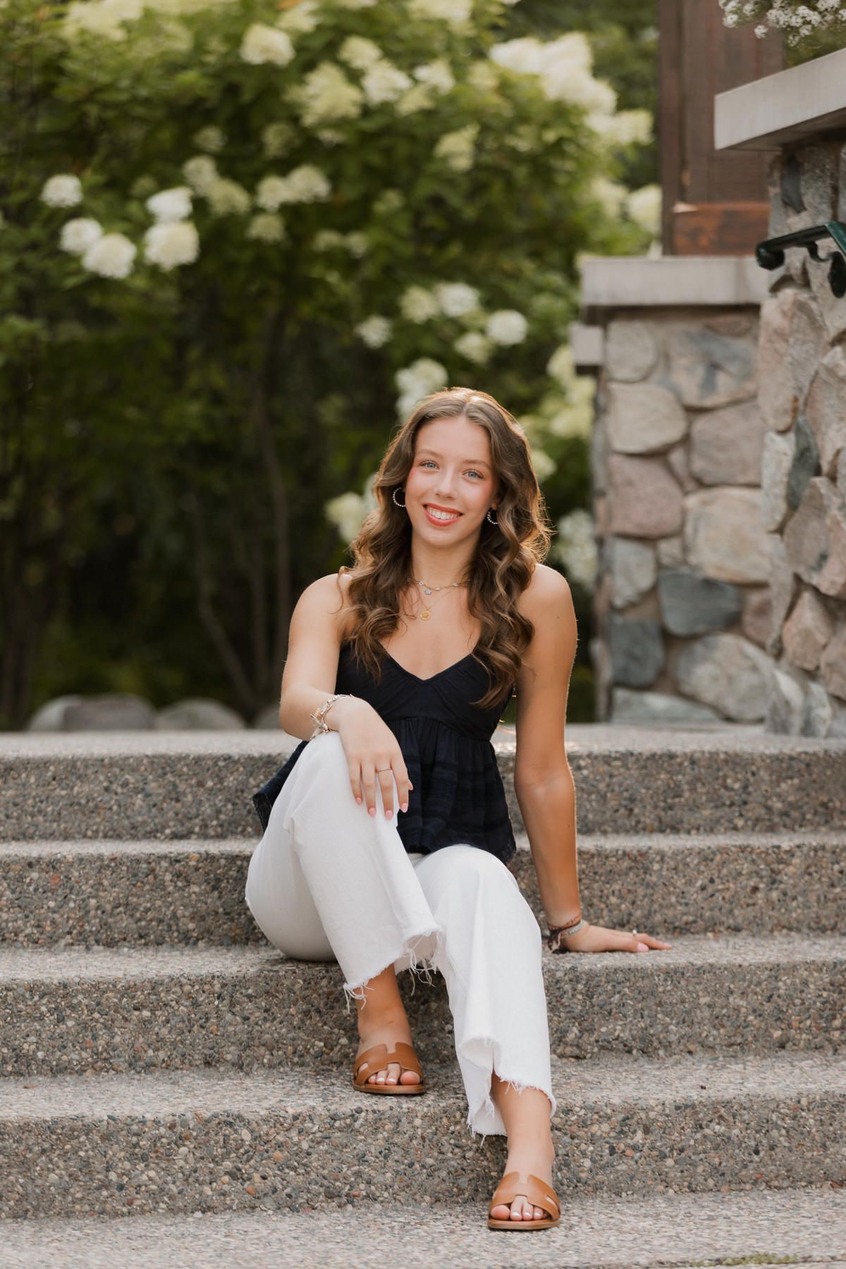 Smiling young woman in black top and white pants sitting on stone steps outdoors.