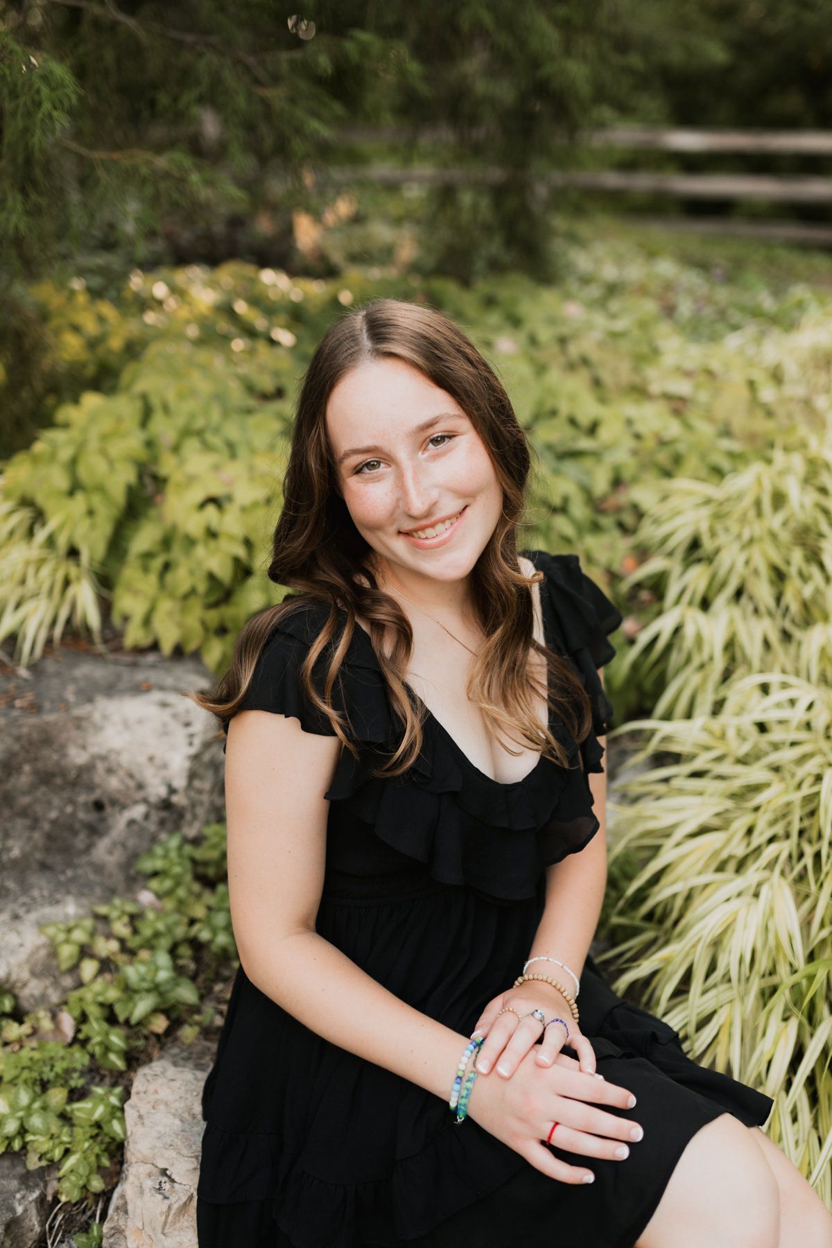 Smiling teenage girl in black ruffled dress sitting on rocks surrounded by lush green garden foliage.