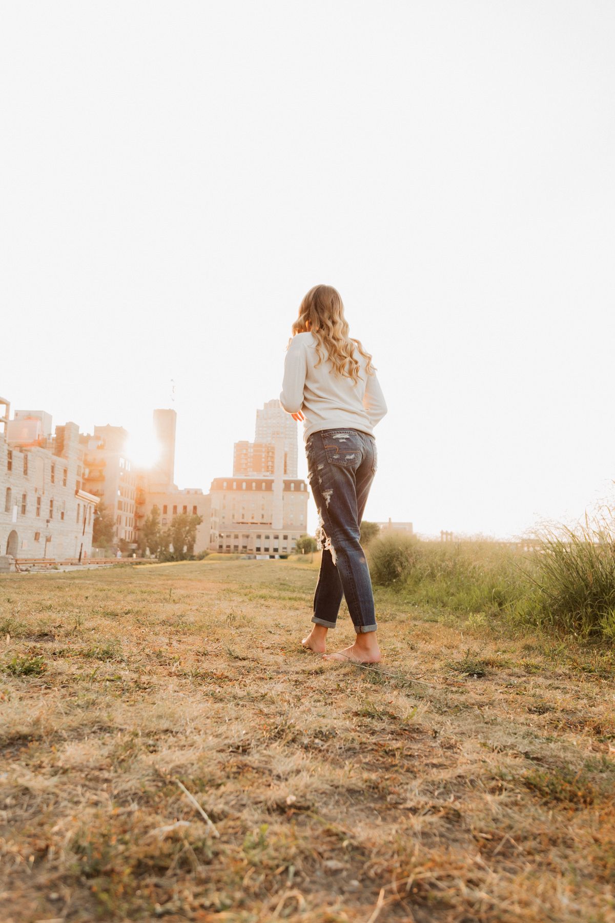 Young woman in jeans and sweater stands in golden sunset light with city buildings behind.