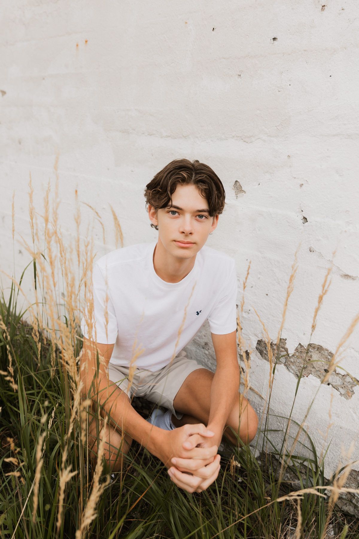 Young man in white t-shirt and shorts sits cross-legged in tall grass against a white weathered wall.