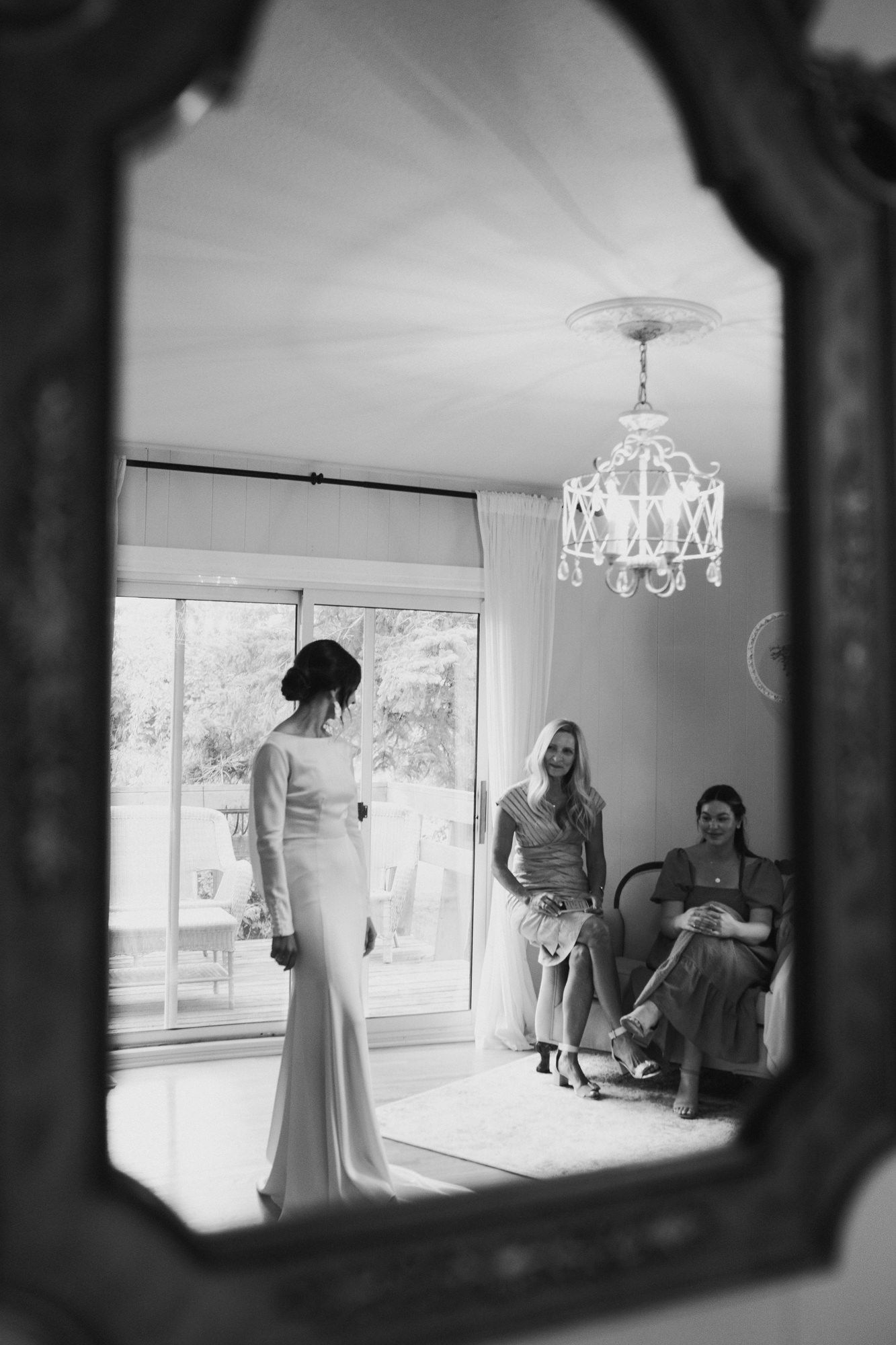 Black and white mirror reflection of a bride in a fitted gown with two seated bridesmaids and chandelier.