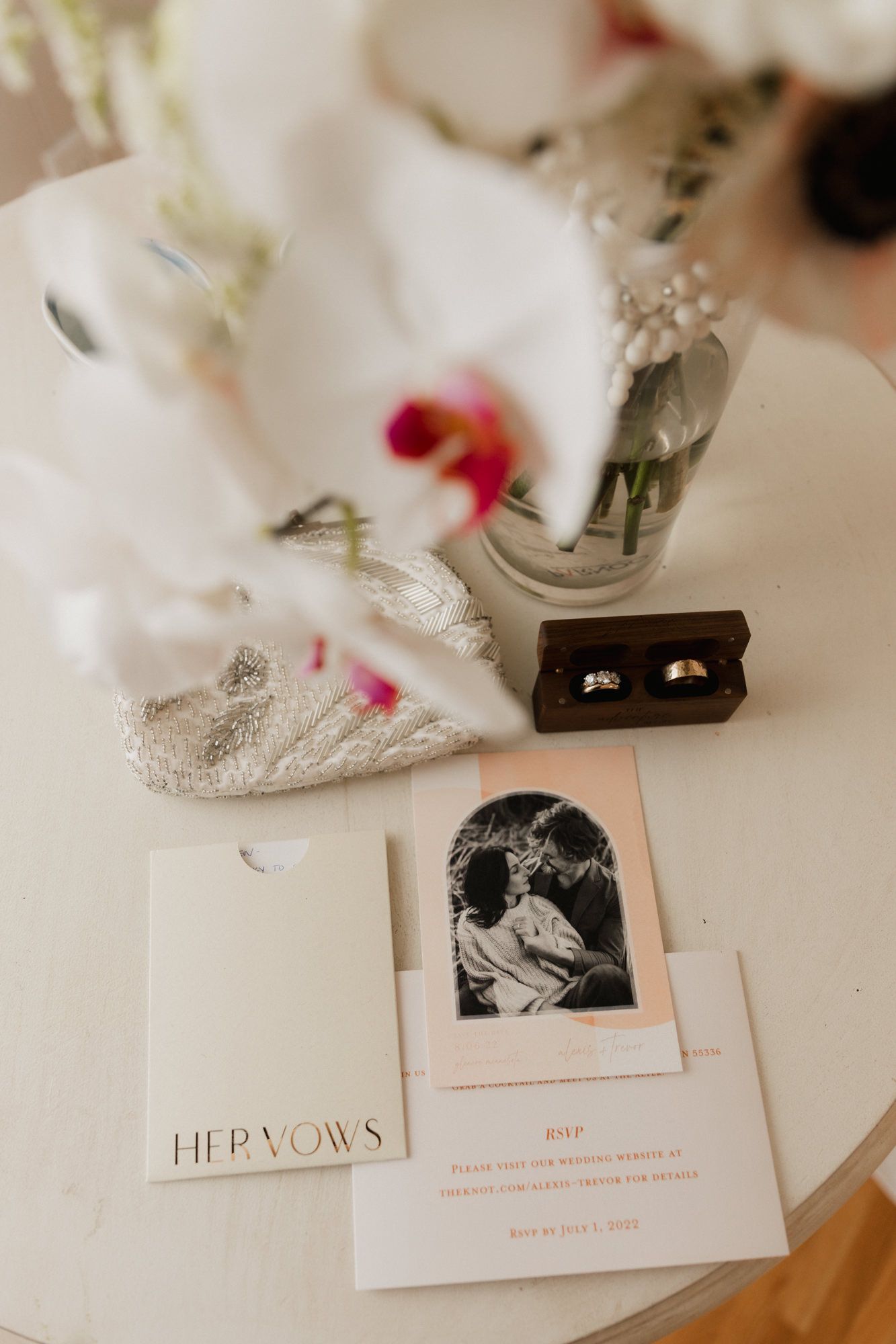Elegant wedding flat lay with vow booklet, RSVP card, engagement photo, gold rings, and floral arrangement on white table.