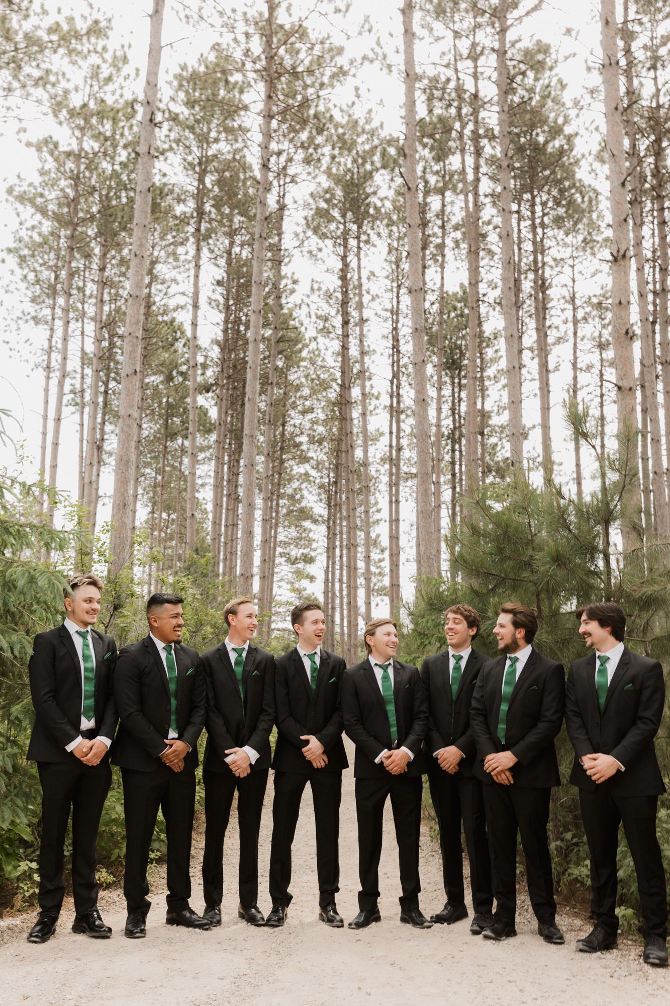 Groomsmen in black suits with green ties standing in a pine forest for a wedding party photo.