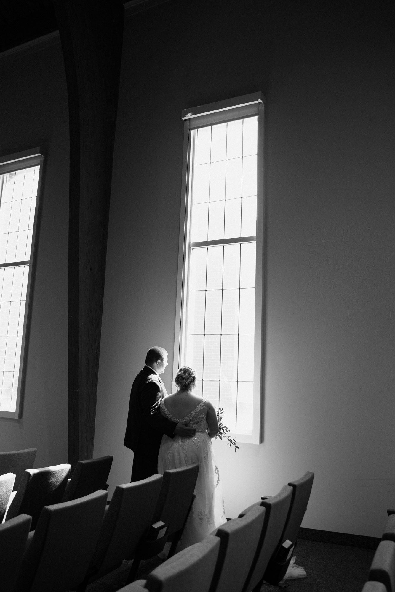 Black and white photo of bride and groom standing before tall chapel windows, holding bouquet.