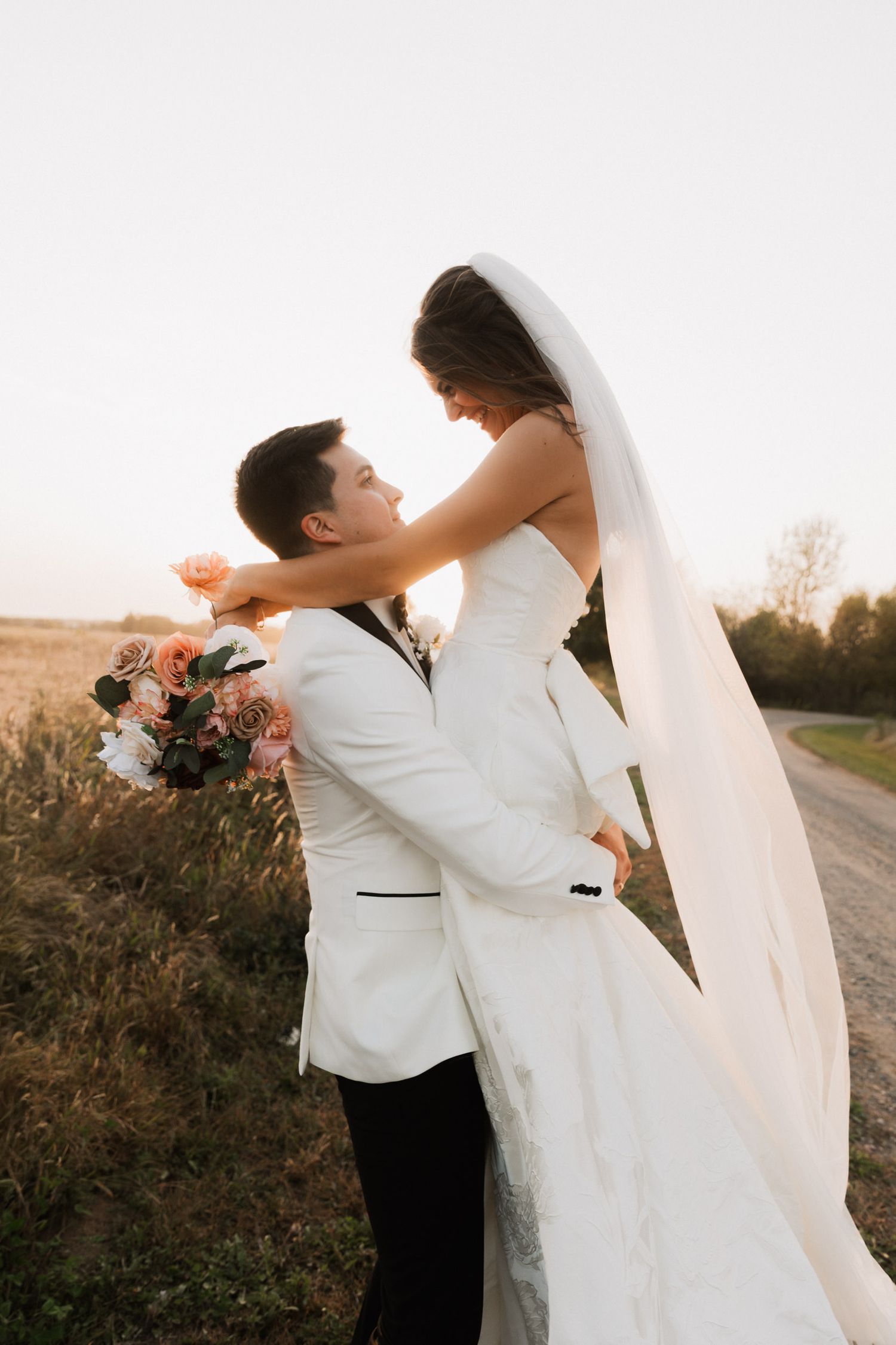 Groom in white tuxedo lifts bride in wedding gown and veil, sharing a romantic moment in a golden field at sunset.