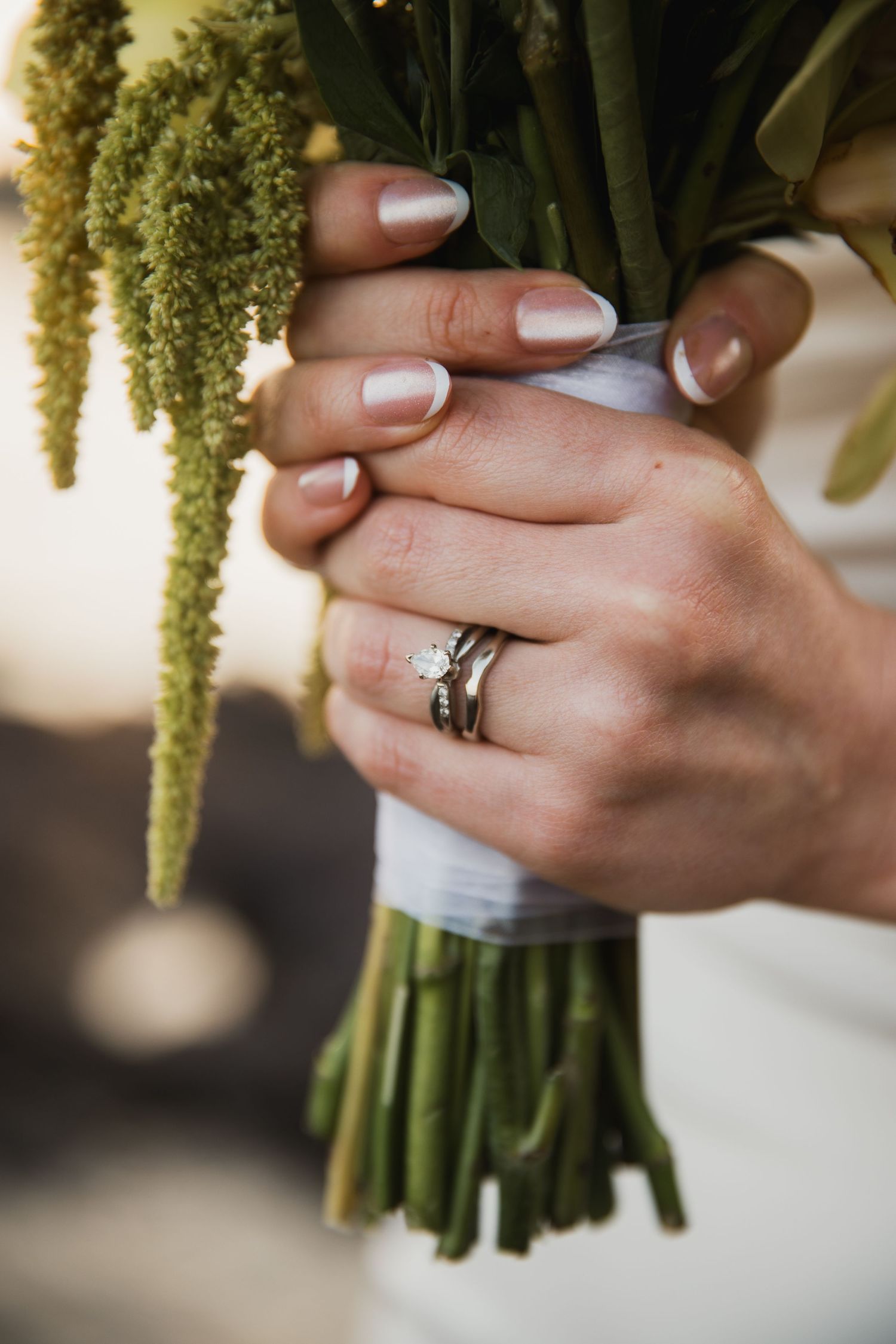 Close-up of bride's hands holding green bouquet, showcasing diamond engagement ring and wedding band with French manicure.