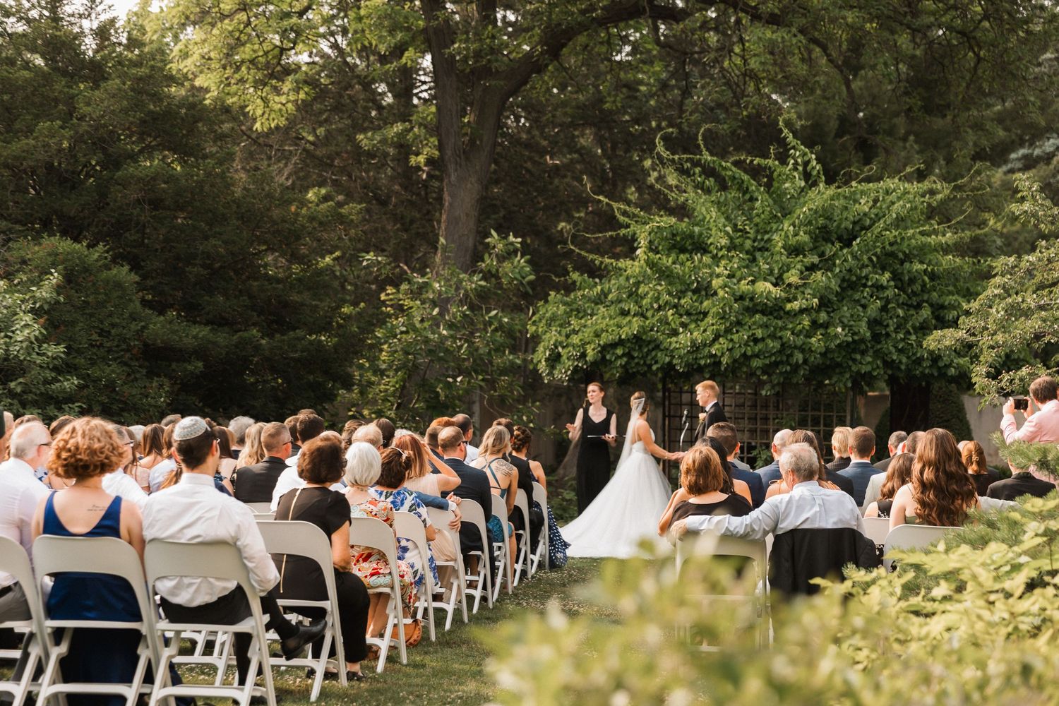 Outdoor wedding ceremony with guests seated on white chairs facing couple at altar surrounded by lush green trees.