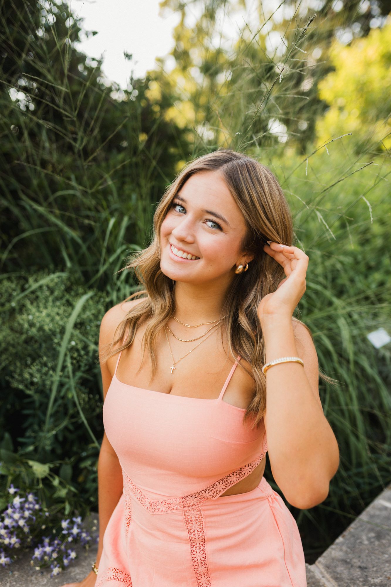 Young woman in pink outfit smiling outdoors surrounded by lush green foliage on a sunny day.