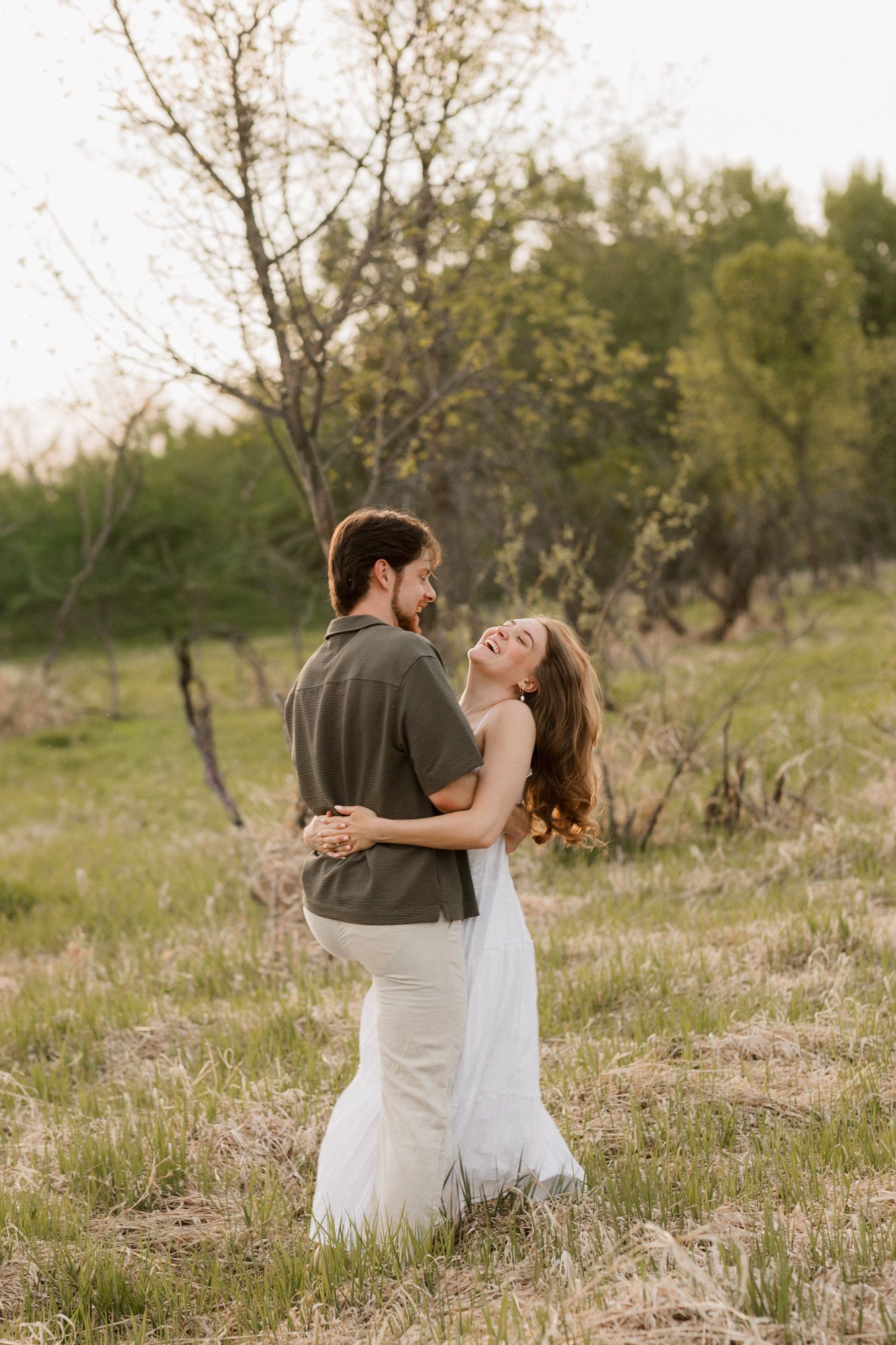 Couple embracing and laughing in a green field, woman in white dress, man in olive shirt, trees in background.