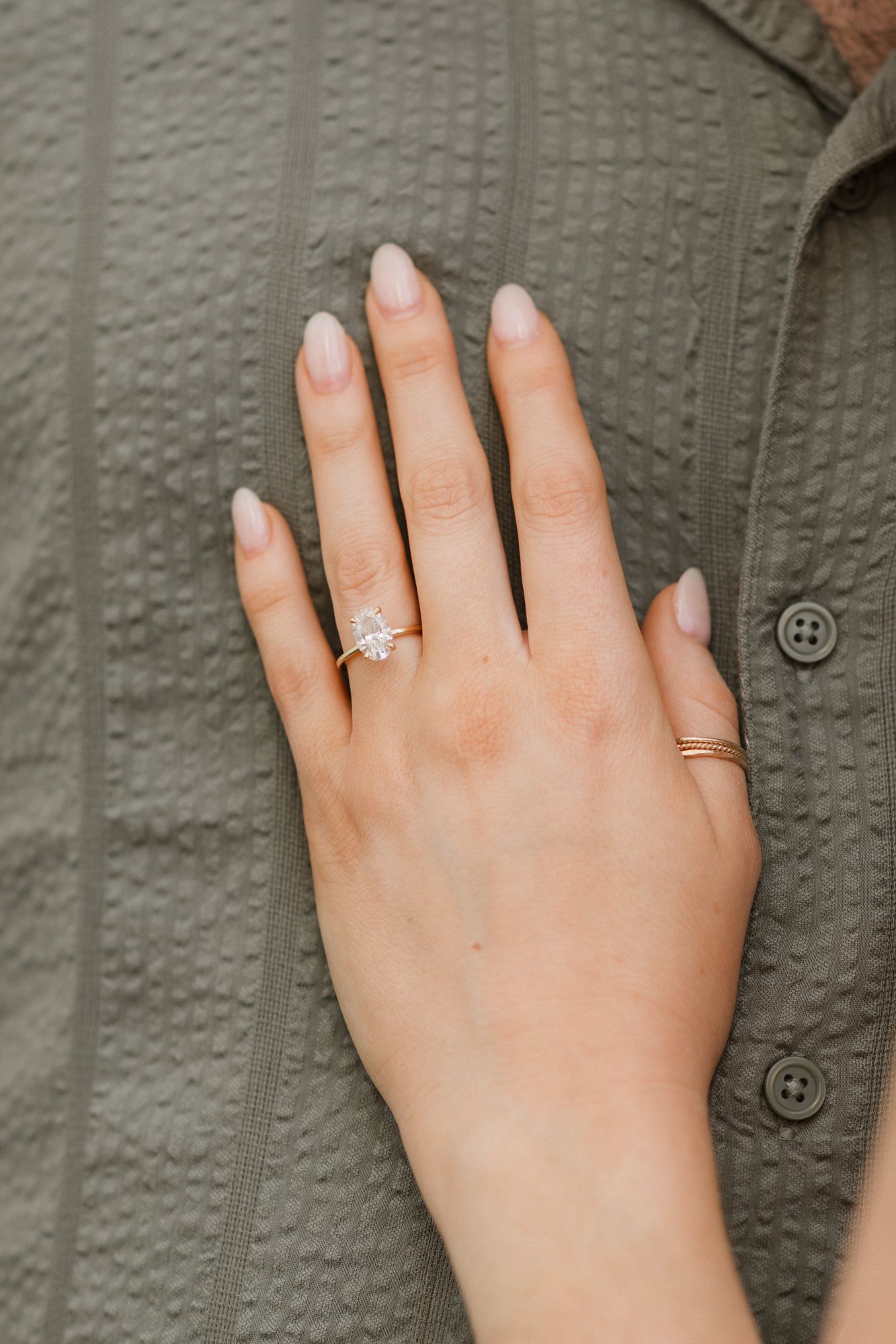 Close-up of a hand wearing a sparkling diamond engagement ring and gold band, resting on an olive green textured shirt.