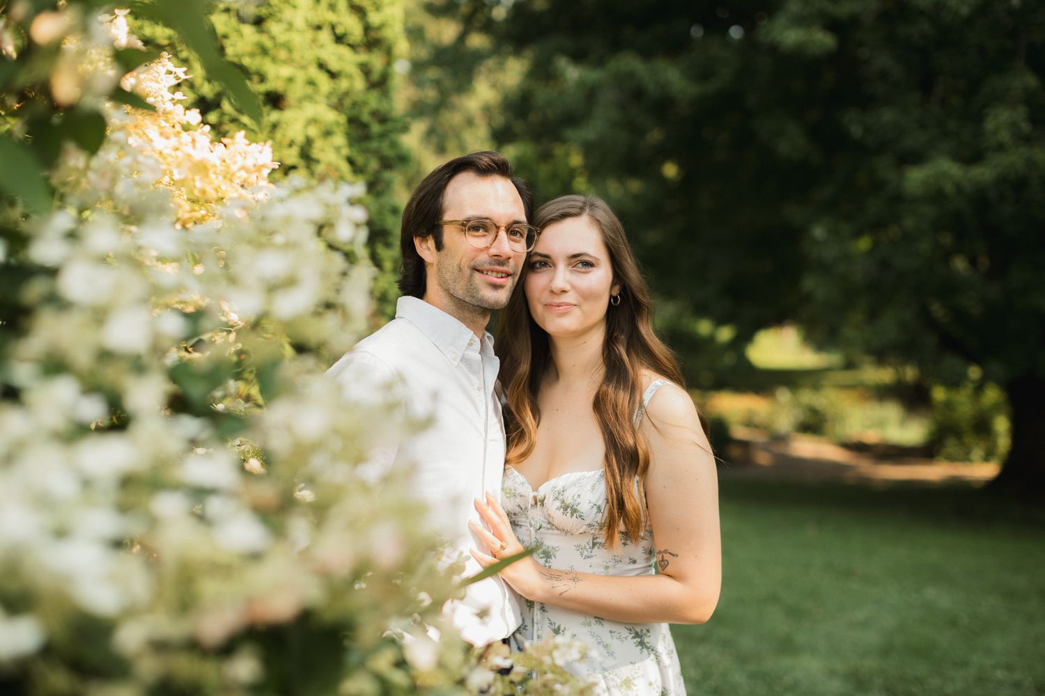 Couple poses together among white blooming flowers in a lush green garden on their wedding day.