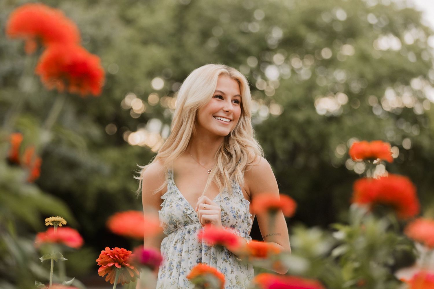 Smiling blonde woman in floral dress surrounded by vibrant red and orange zinnia flowers in a lush garden.