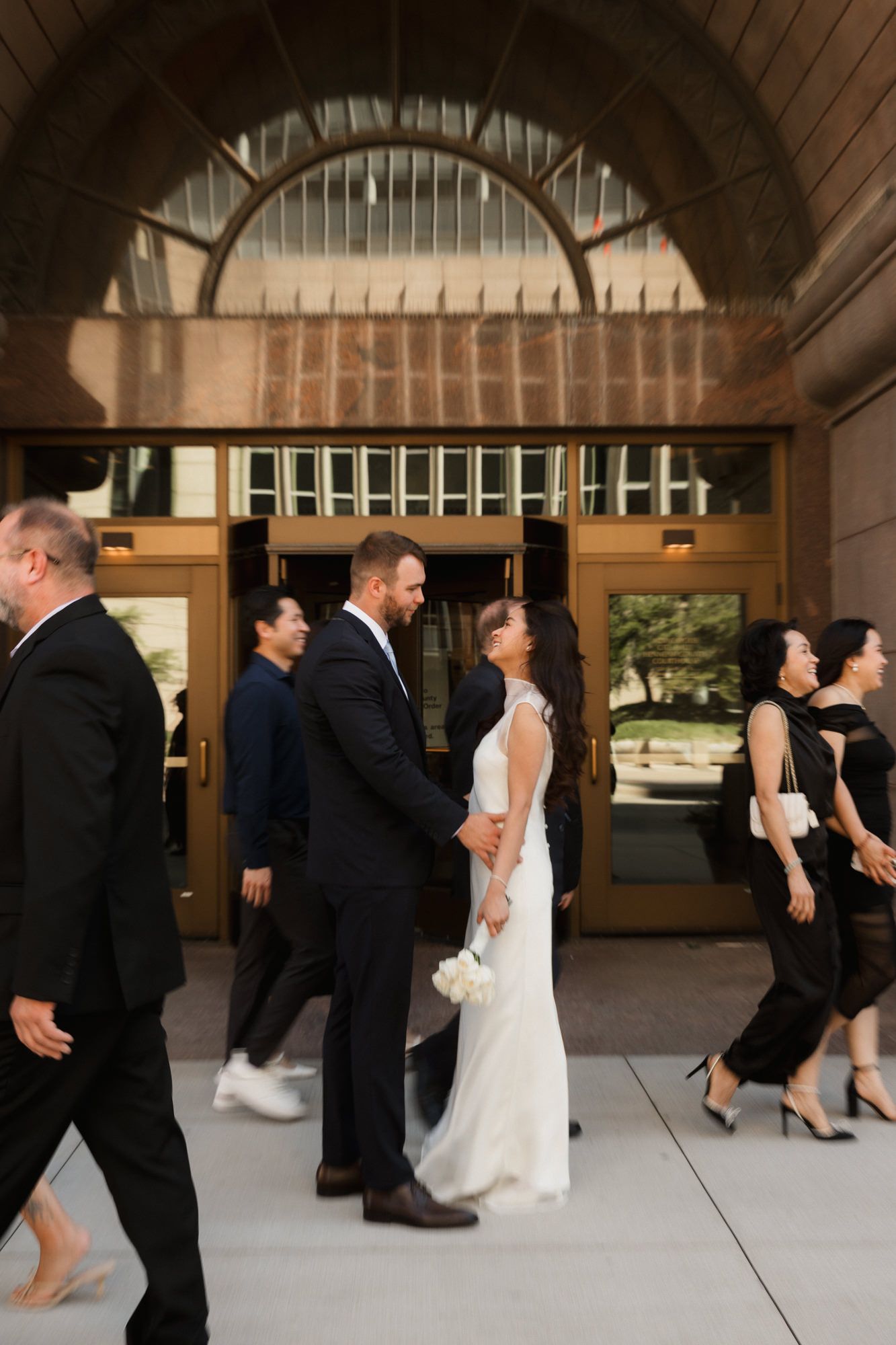 Bride and groom hold hands outside elegant building entrance surrounded by wedding guests in formal attire.