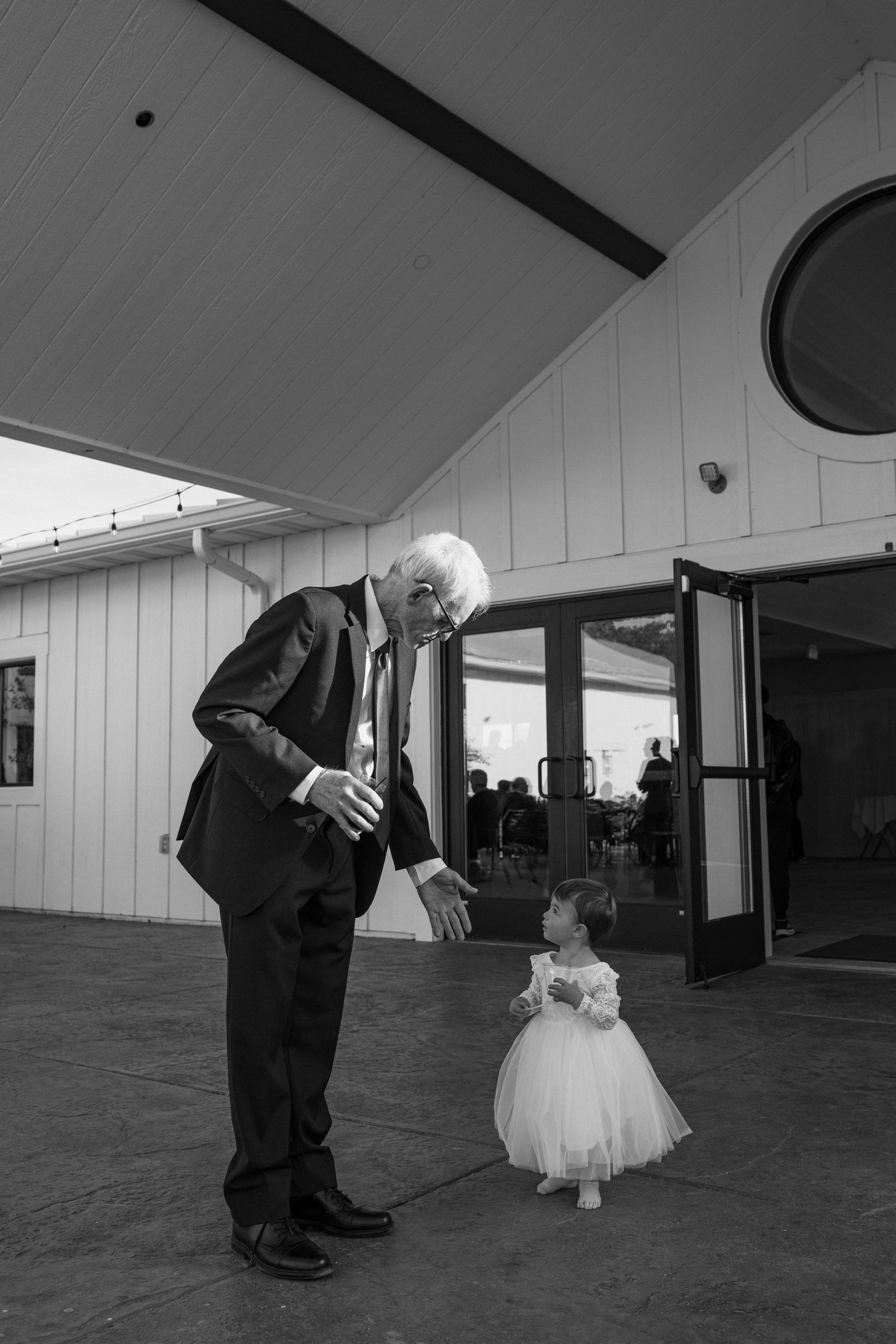 Elderly man in a suit reaches toward a toddler in a tulle flower girl dress outside a rustic wedding venue in black and white.