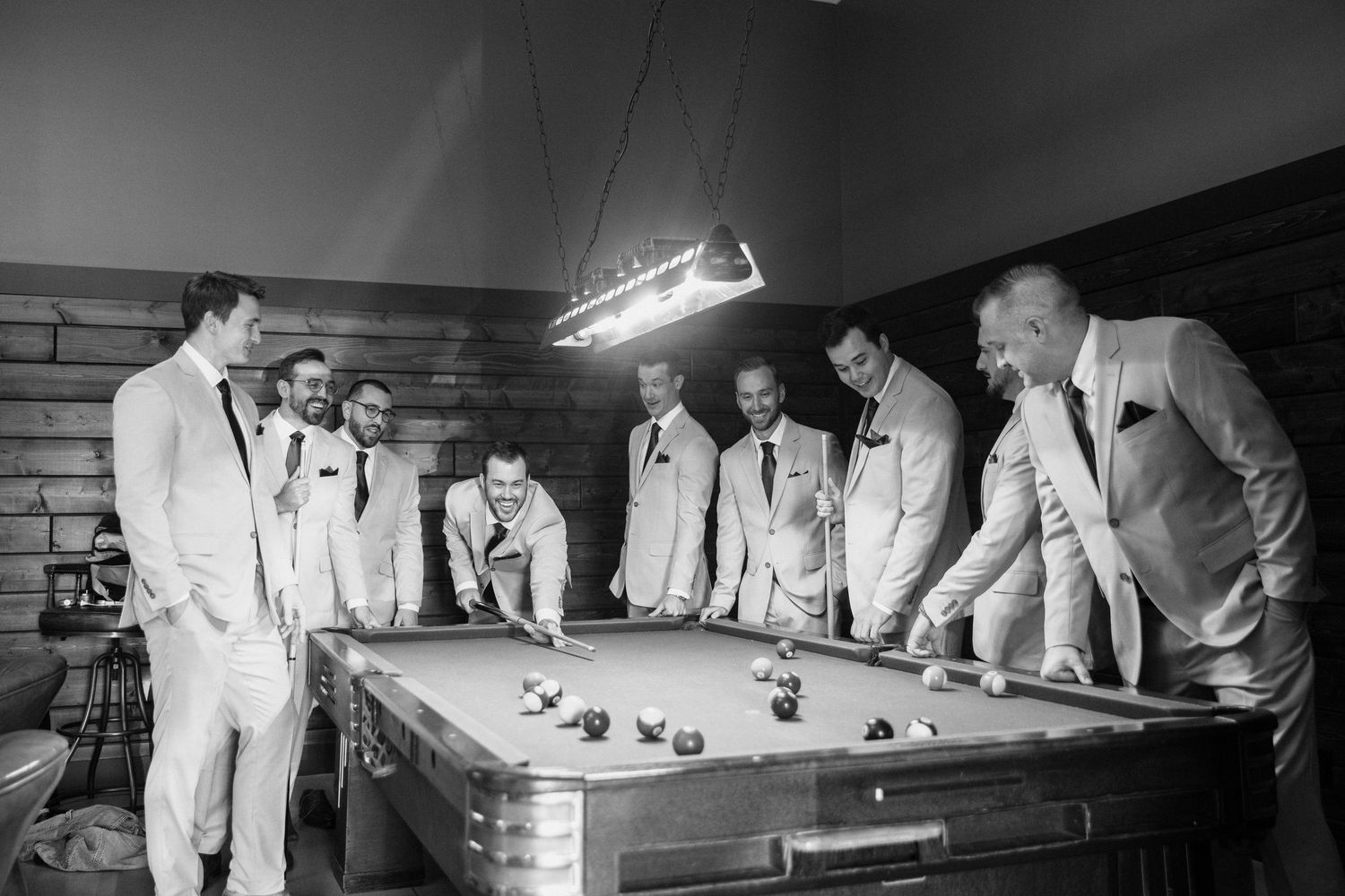 Black and white photo of groomsmen in suits gathered around a pool table, laughing and playing billiards under a hanging light.