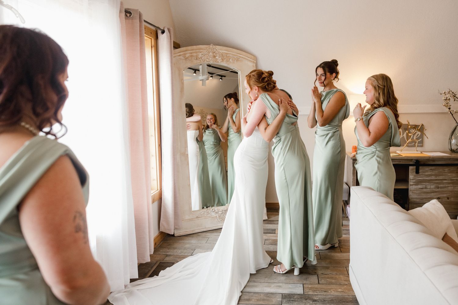 Bride in white gown views reflection in mirror surrounded by bridesmaids in sage green dresses during wedding preparations.